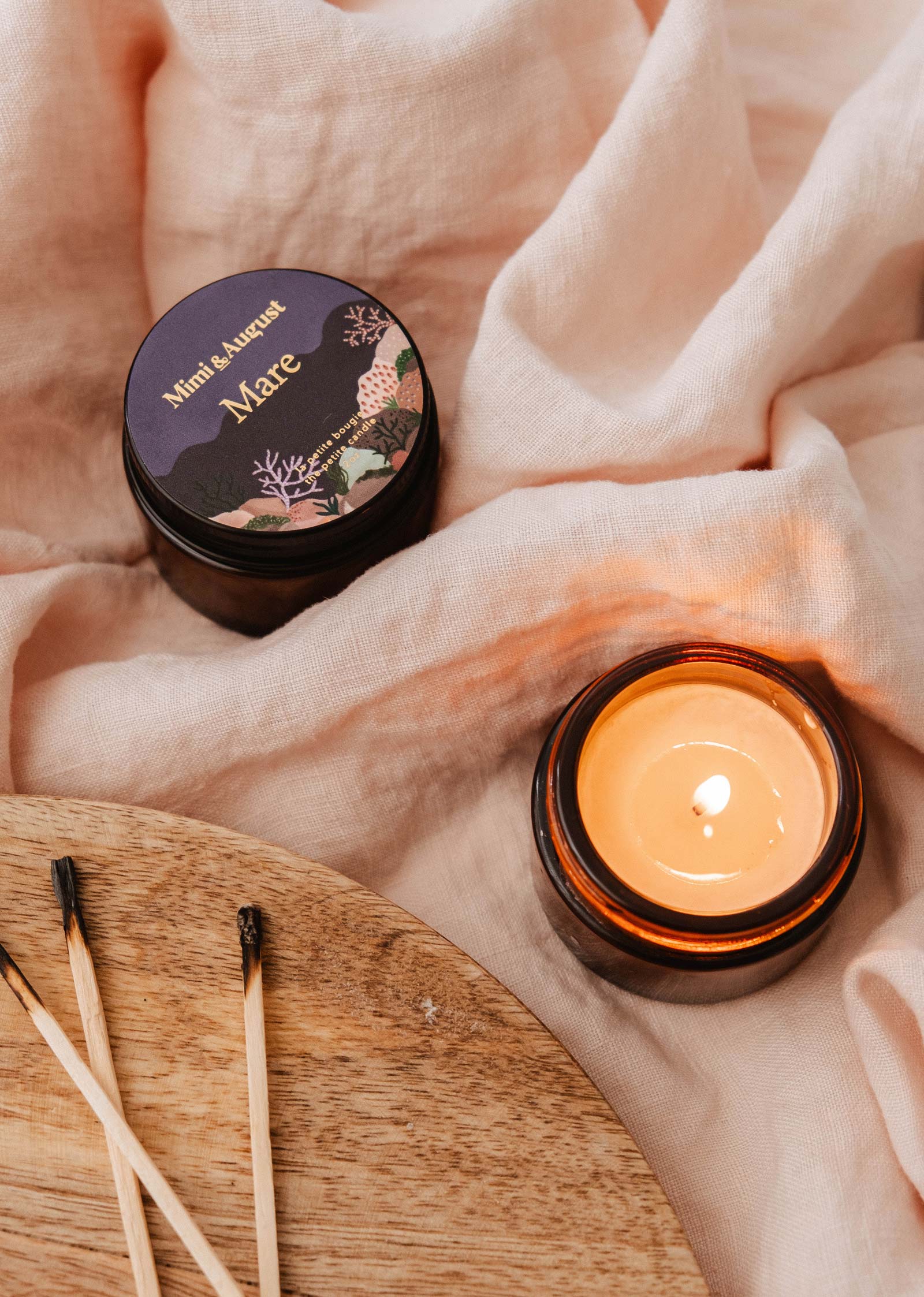 The Mimi & August Mare - 2 oz reusable candle in a brown glass jar sits on soft beige fabric, evoking a calming mood, with burnt matchsticks scattered nearby.