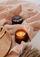 A Mare - 2 oz candle from Mimi & August in amber glass burns beside its matching jar on soft pink fabric with wooden matches and a magazine, filling the air with a calming Ocean scent.
