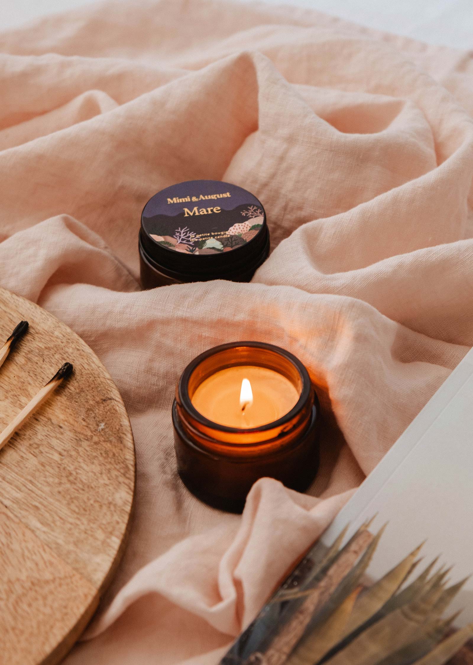 A Mare - 2 oz candle from Mimi & August in amber glass burns beside its matching jar on soft pink fabric with wooden matches and a magazine, filling the air with a calming Ocean scent.