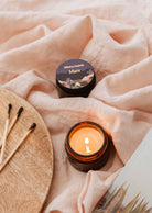 The Mare - 2 oz reusable candle by Mimi & August, in a small brown jar, rests on soft pink fabric next to a closed jar, matches, and wood, creating a calming atmosphere.
