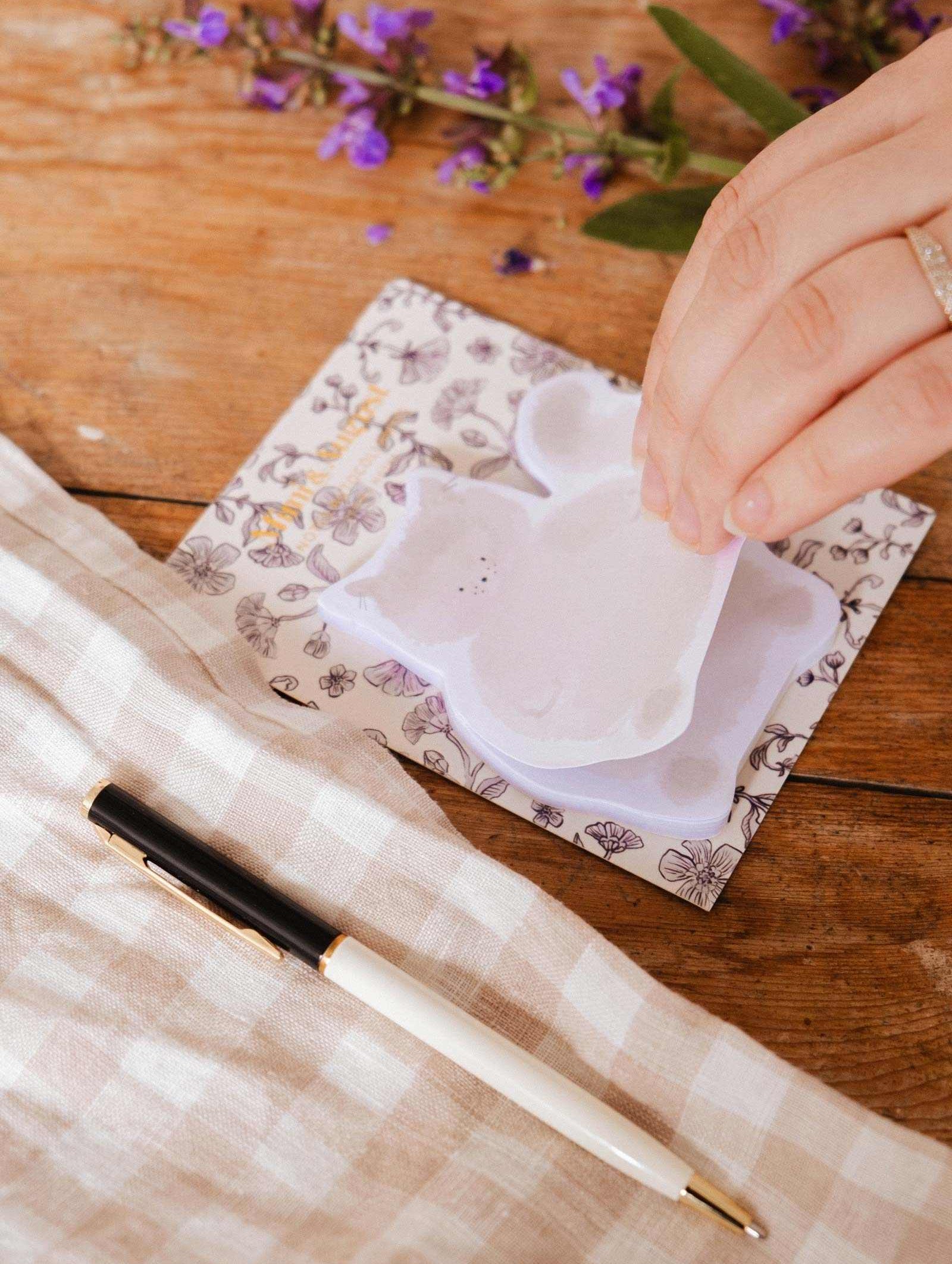 A hand lifts a cat-shaped Meow - Sticky Note by Mimi & August from a floral notepad on a wooden table with a black and white pen, beige checkered cloth, and purple flowers—perfect for cute stationery fans.