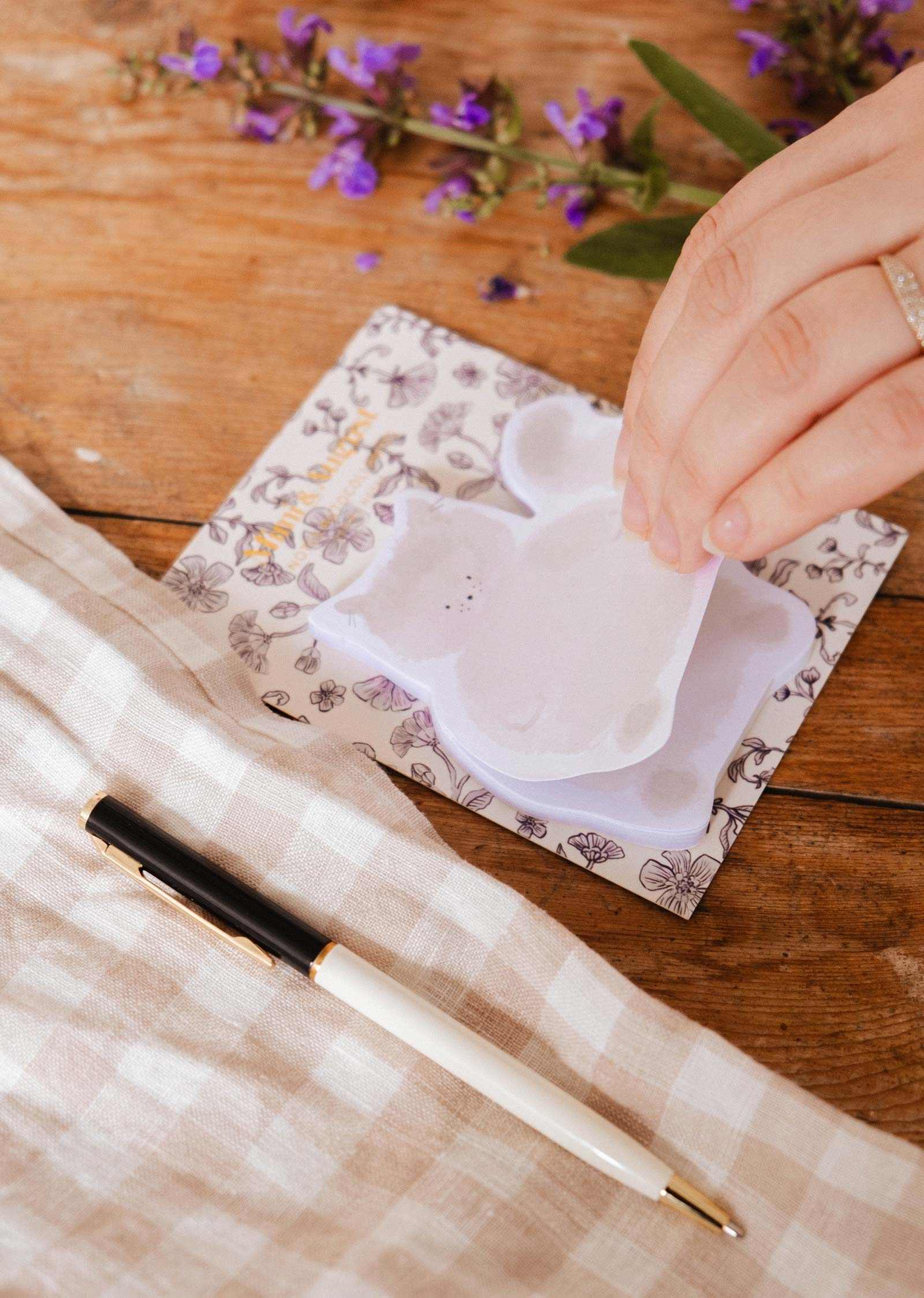 A hand lifts a cat-shaped Meow - Sticky Note by Mimi & August from a floral notepad on a wooden table with a black and white pen, beige checkered cloth, and purple flowers—perfect for cute stationery fans.
