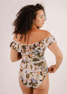 A person with curly hair stands sideways, donning a Mimi & August Milano Mariposa One-Piece Swimsuit showcasing a stunning butterfly and flower print.