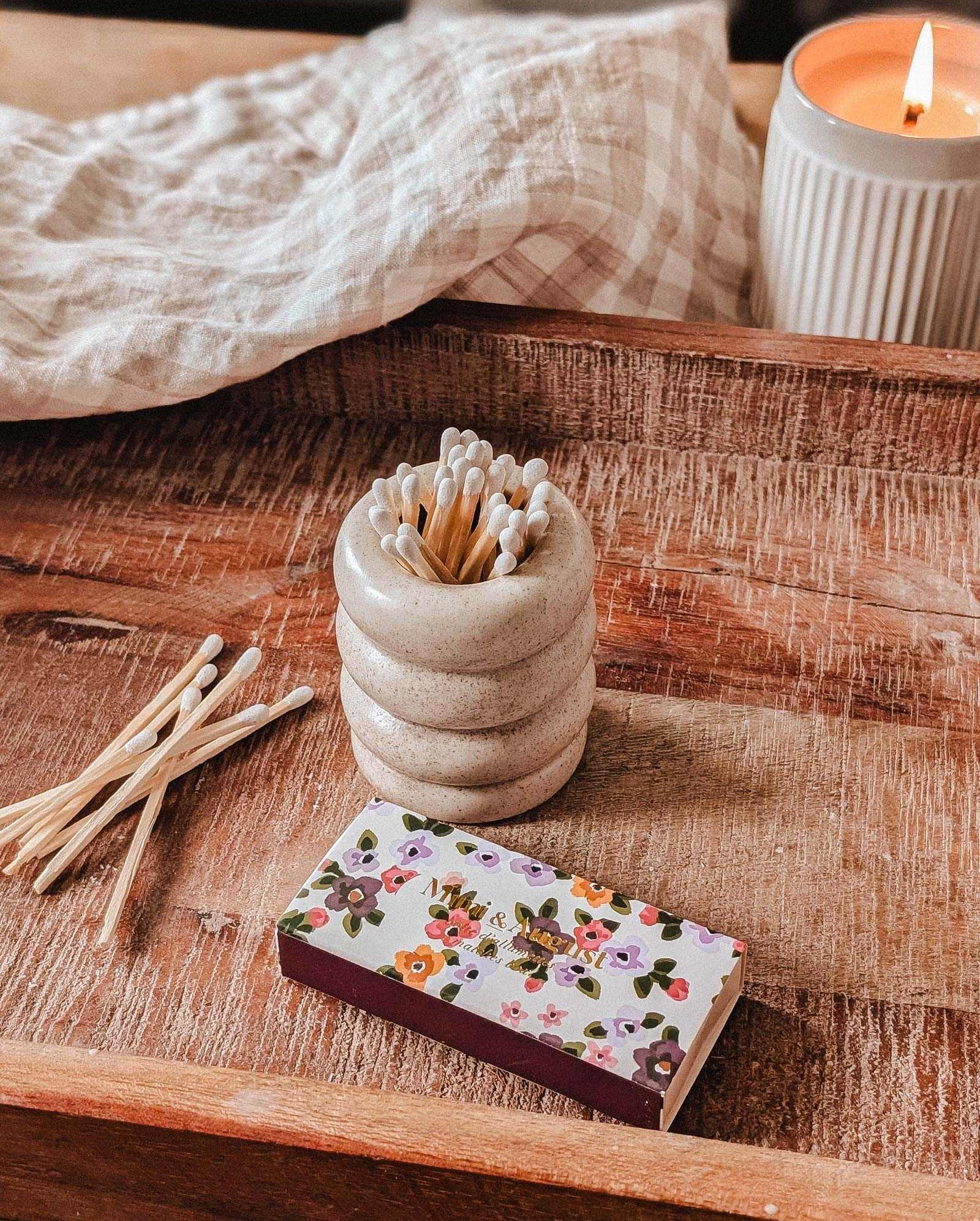 A wooden tray holds a lit candle, a ceramic match holder with decorative matches, several loose matches, and the Mimi Floral Matchbox - 30 Sticks by Mimi & August. A checked cloth is draped in the background.
