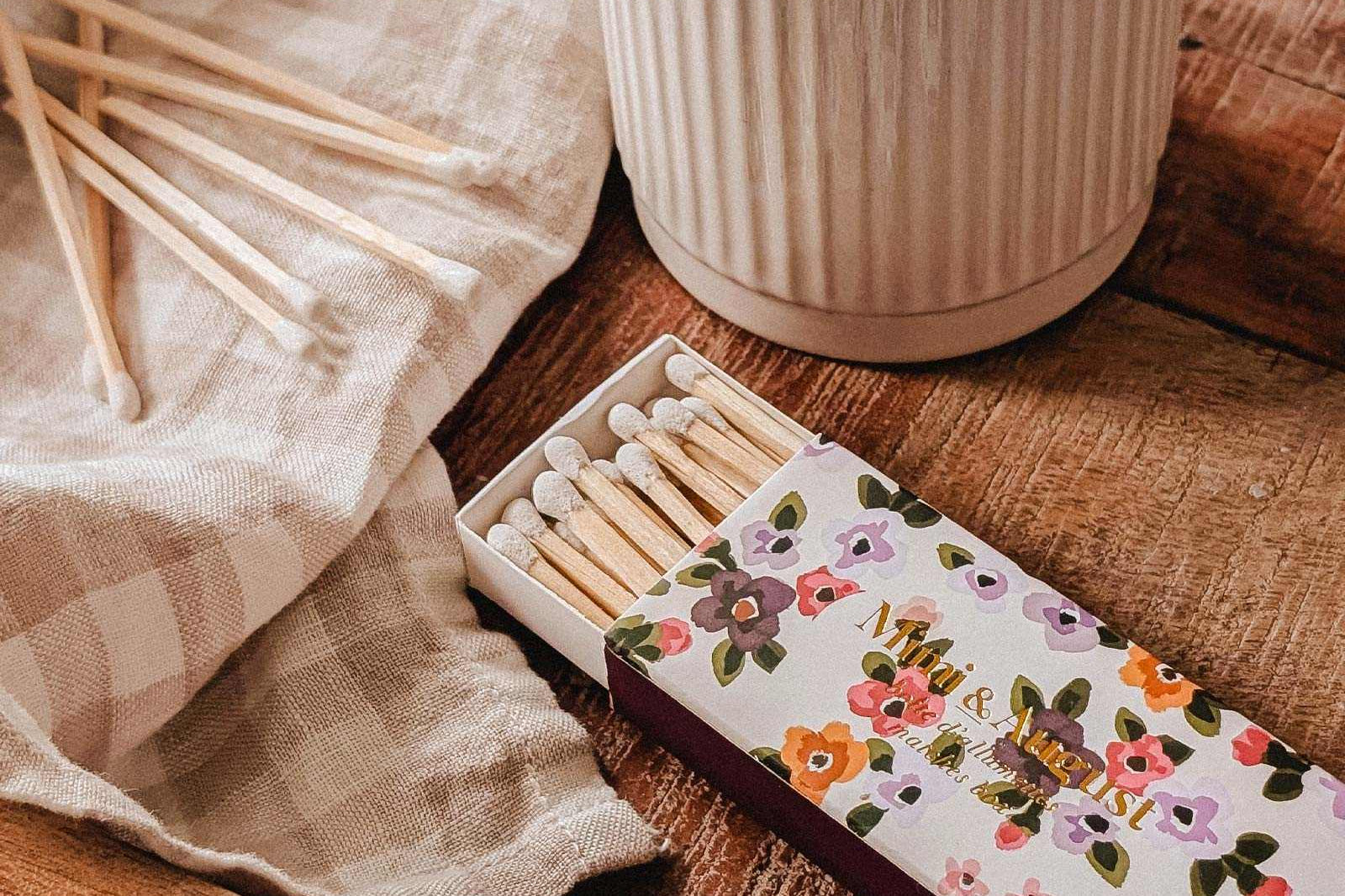 A lit candle in a ceramic holder rests on a wooden tray alongside a Mimi & August Mimi Floral Matchbox (30 sticks), several loose matches, and a checkered cloth.