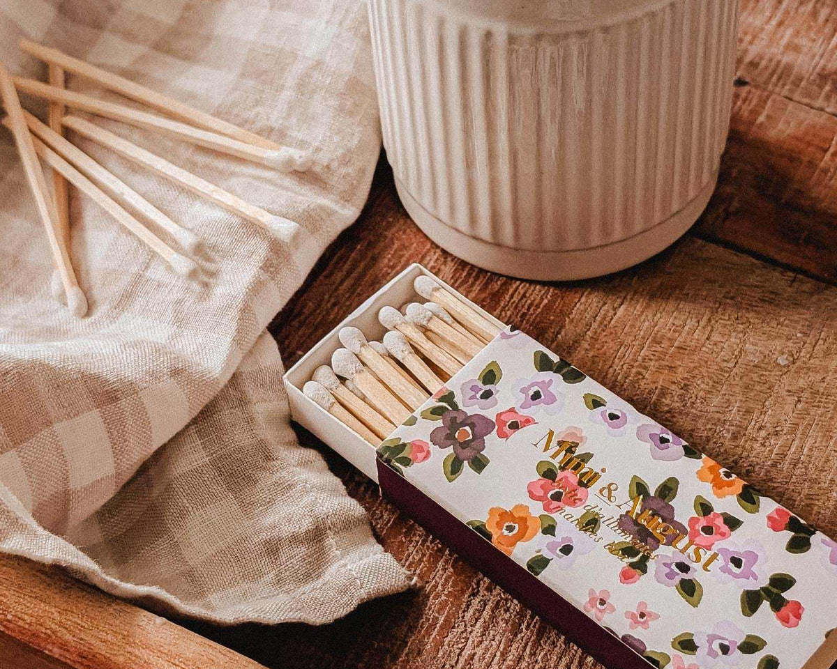 A lit candle in a ceramic holder rests on a wooden tray alongside a Mimi & August Mimi Floral Matchbox (30 sticks), several loose matches, and a checkered cloth.