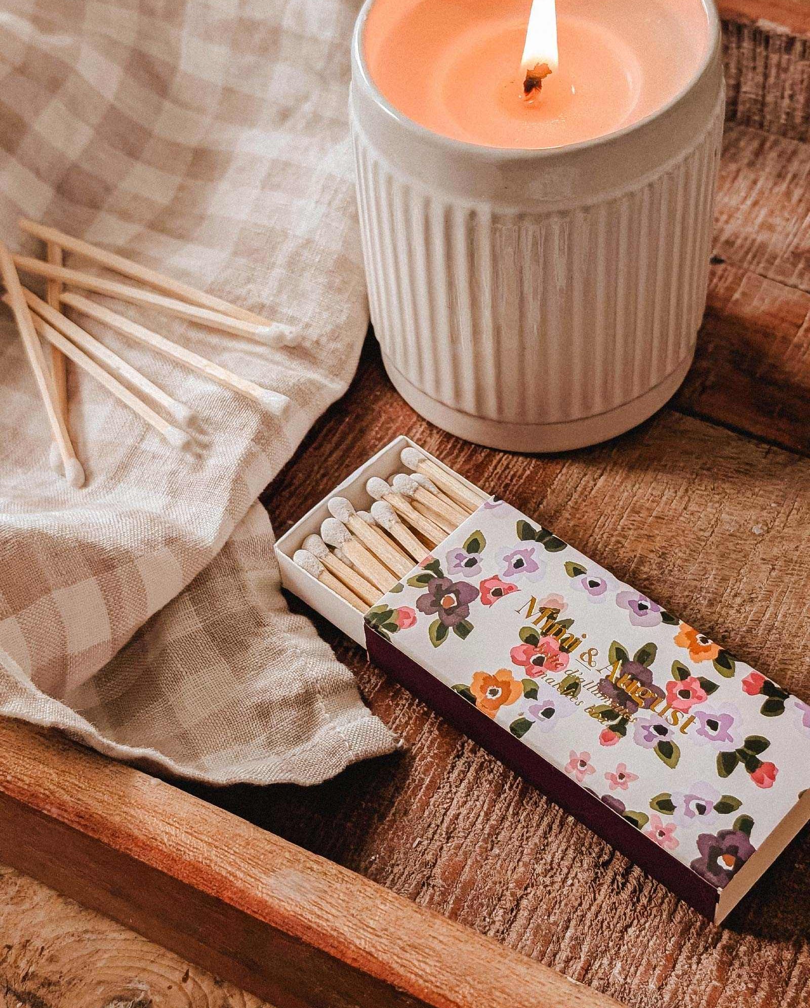 A lit candle in a ceramic holder rests on a wooden tray alongside a Mimi & August Mimi Floral Matchbox (30 sticks), several loose matches, and a checkered cloth.