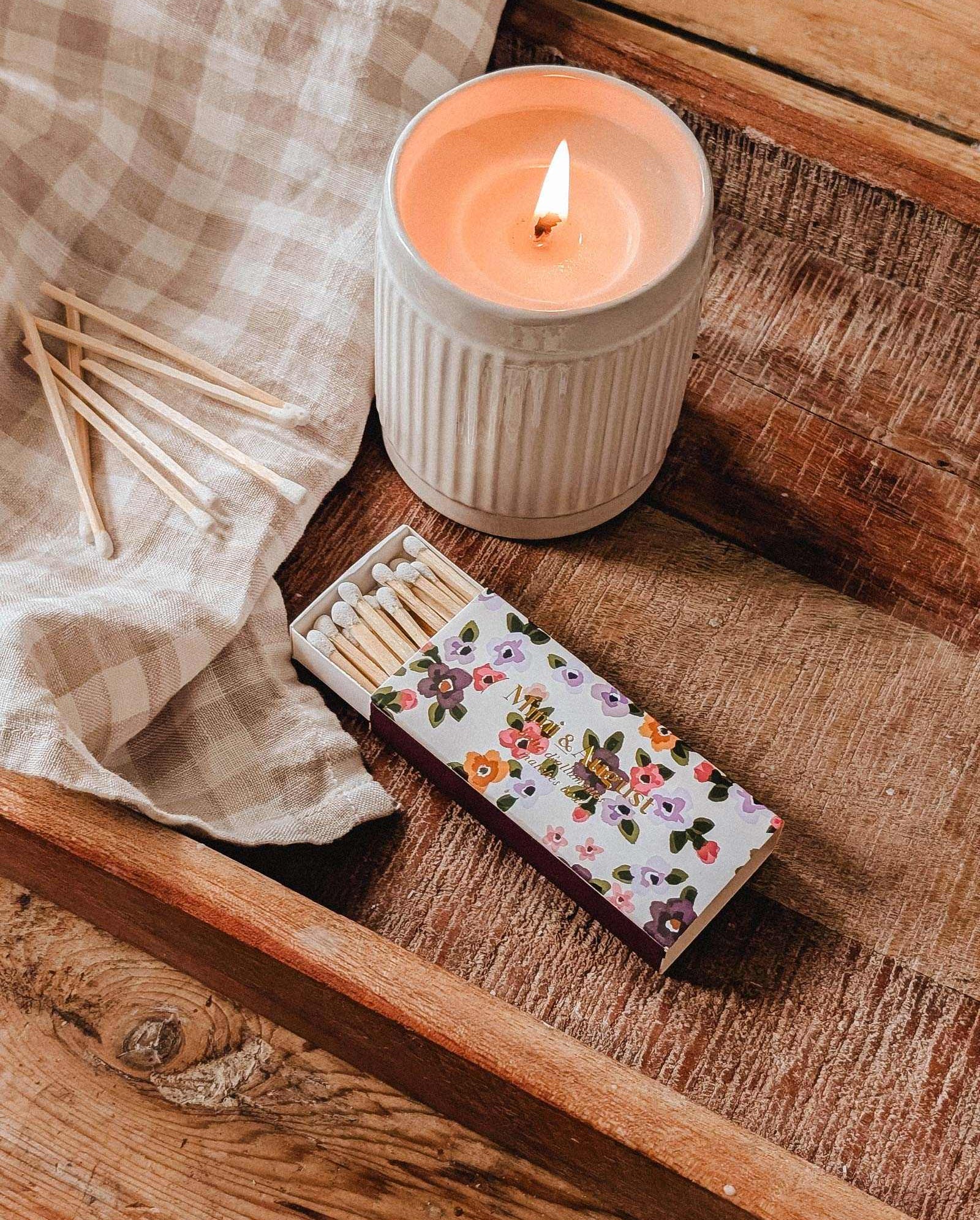 A lit candle in a ribbed white holder sits on a wooden tray beside the Mimi Floral Matchbox - 30 Sticks by Mimi & August, decorative matches, and a beige checkered cloth.