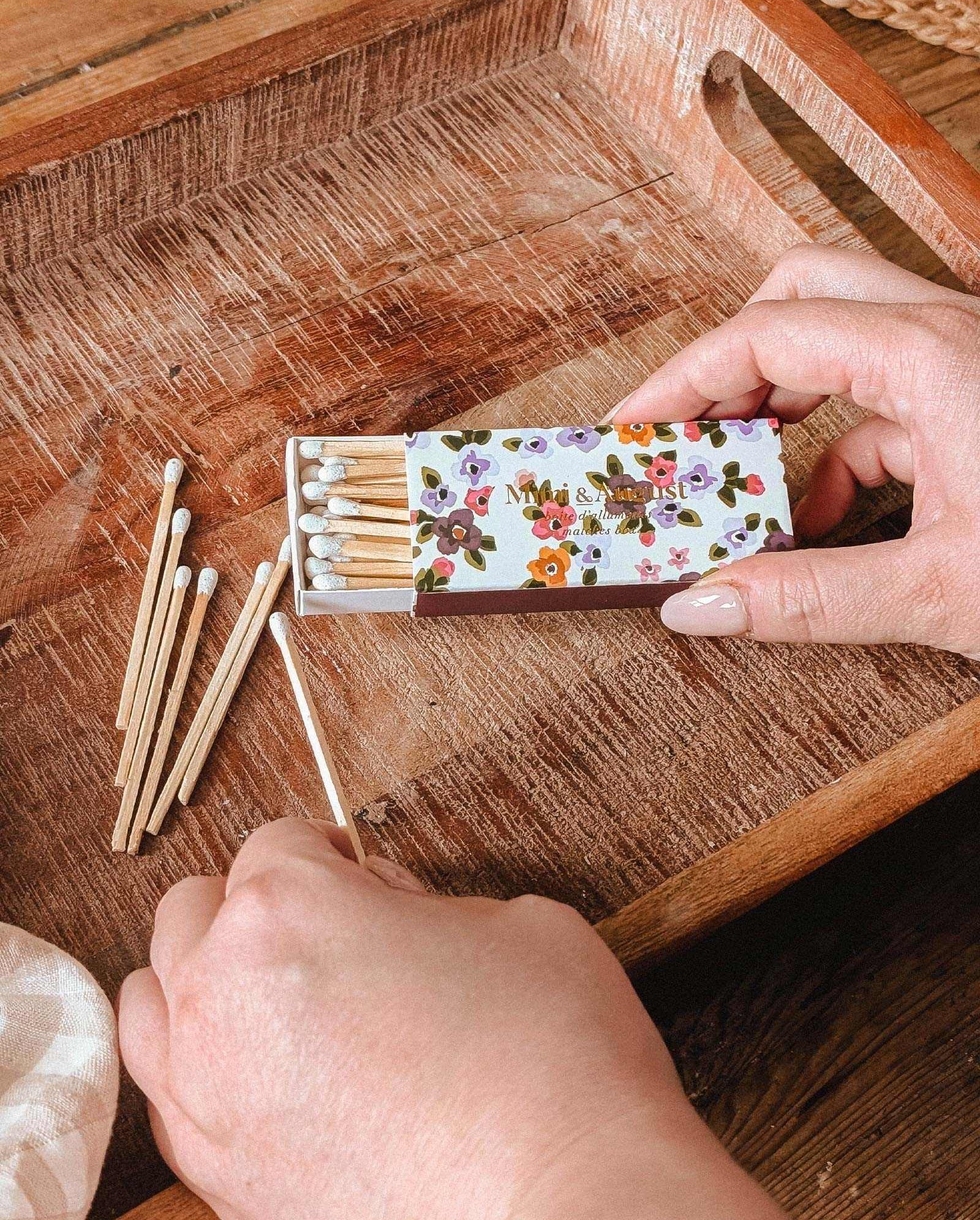 A person holds the Mimi & August Mimi Floral Matchbox - 30 Sticks and takes out a decorative, eco-friendly match, with several more scattered on a wooden tray.