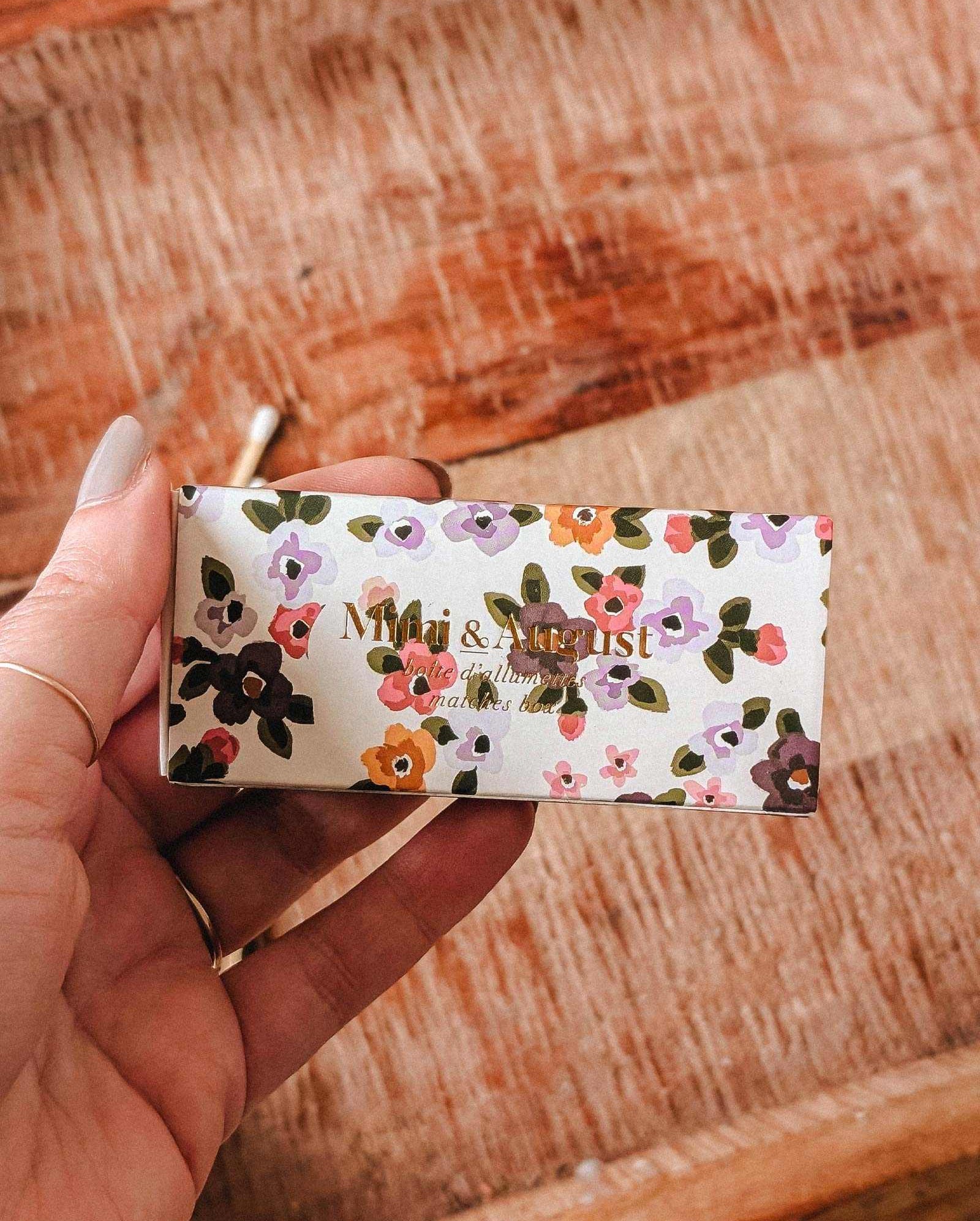 A hand holds a rectangular Mimi & August “Mimi Floral Matchbox - 30 Sticks” over a wooden surface, displaying its decorative matches inside.