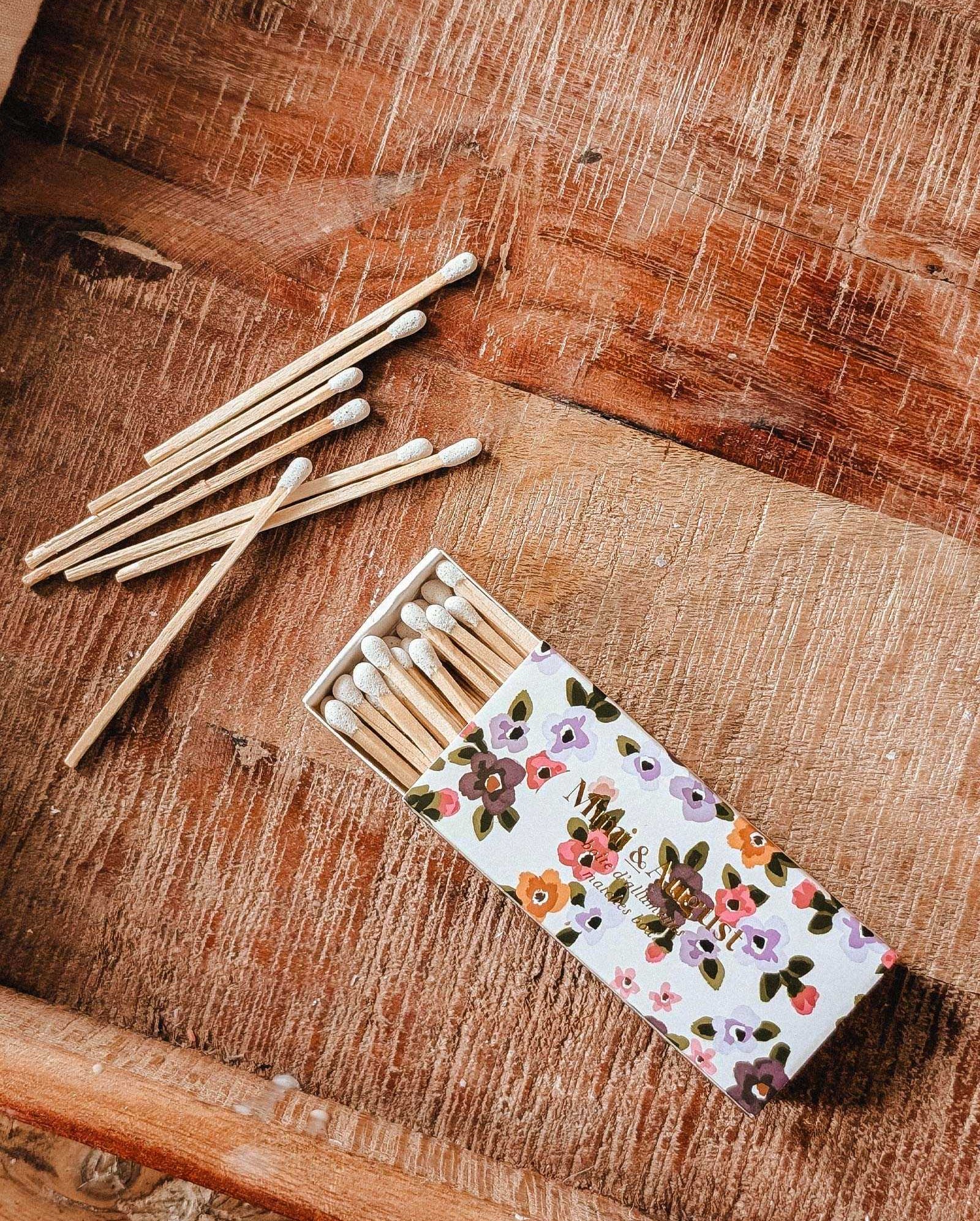 The Mimi & August Mimi Floral Matchbox - 30 Sticks, filled with eco-friendly matches, sits on a wooden surface with several matches scattered nearby.