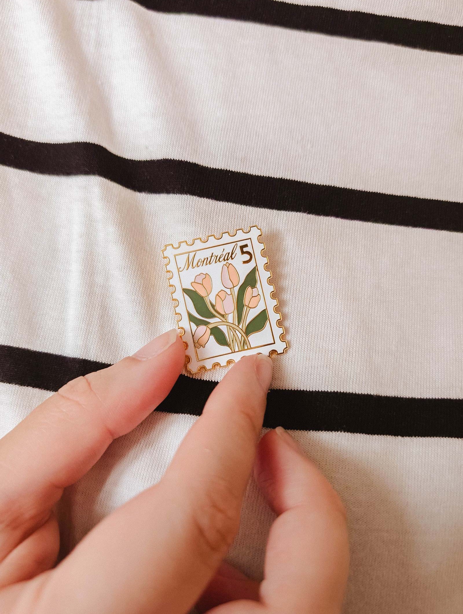 A hand holds the Montréal Stamp Enamel Pin by Mimi & August—an enamel pin styled like a postage stamp with tulips and "Montreal 5" text—set against a black and white striped fabric background. Perfect for flower lovers.