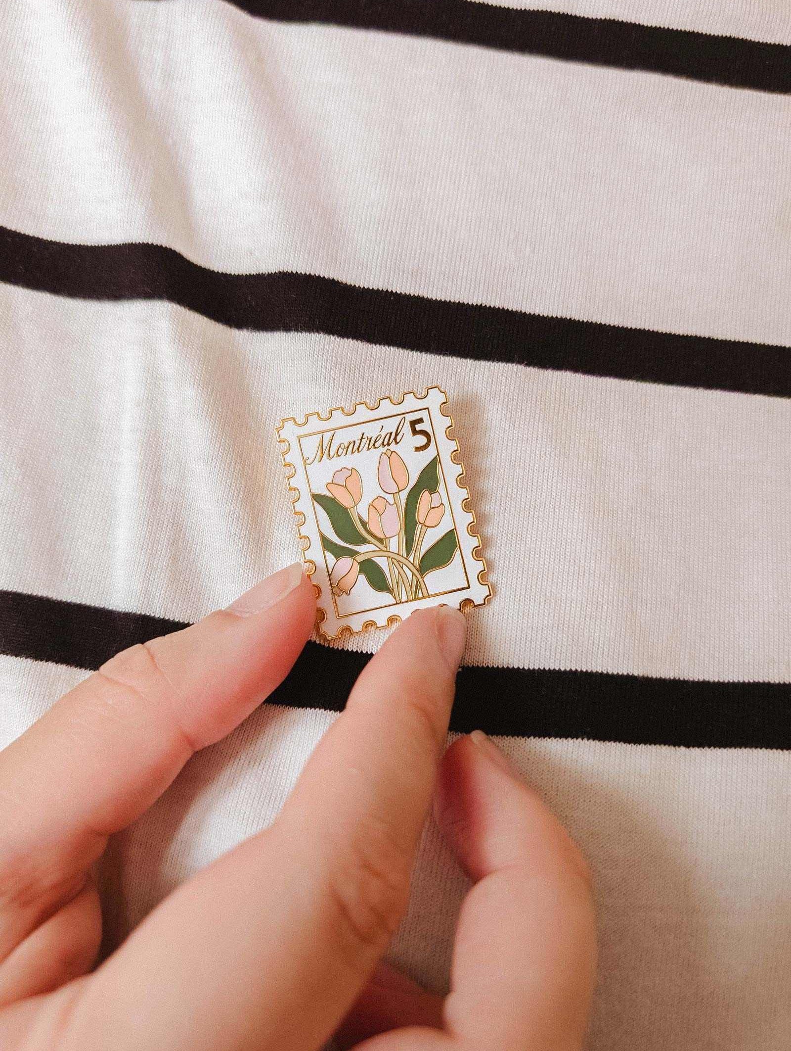 A hand holds the Montréal Stamp Enamel Pin by Mimi & August—an enamel pin styled like a postage stamp with tulips and "Montreal 5" text—set against a black and white striped fabric background. Perfect for flower lovers.