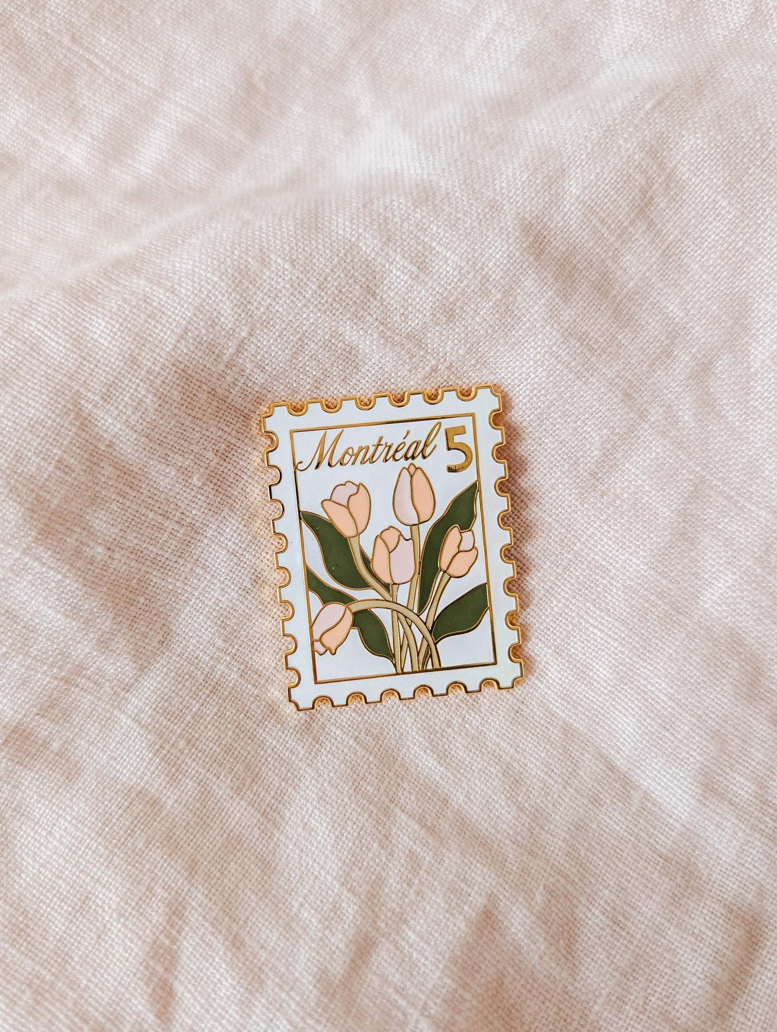 The Montréal Stamp Enamel Pin by Mimi & August is a tulip-themed, stamp-shaped enamel pin with “Montréal 5” text—a charming accessory for flower lovers. Shown on light fabric.