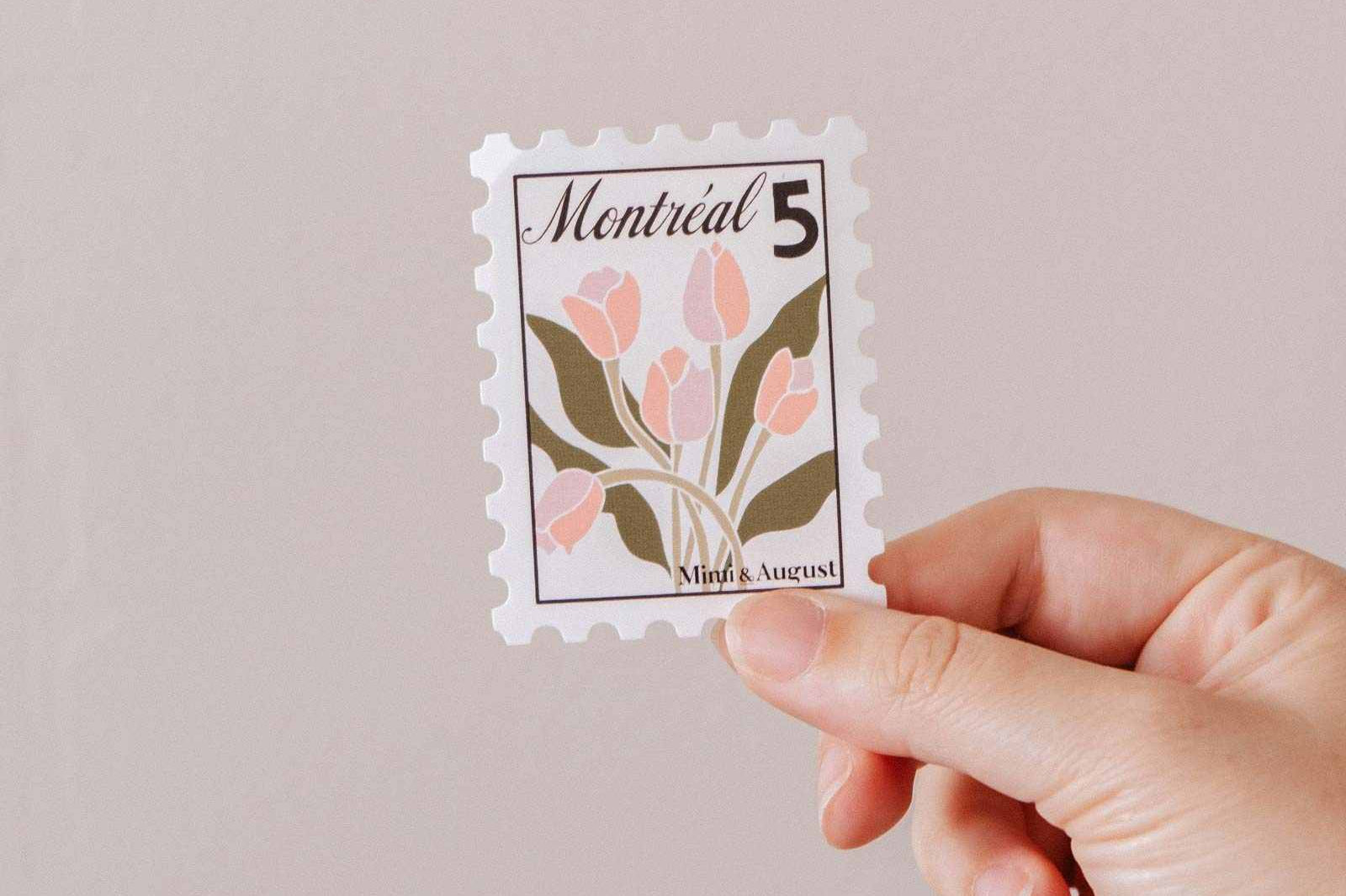 A hand holds the Montréal Stamp - Vinyl Sticker by Mimi & August, featuring pink flowers, green leaves, and "Montréal 5" at the top against a plain background.