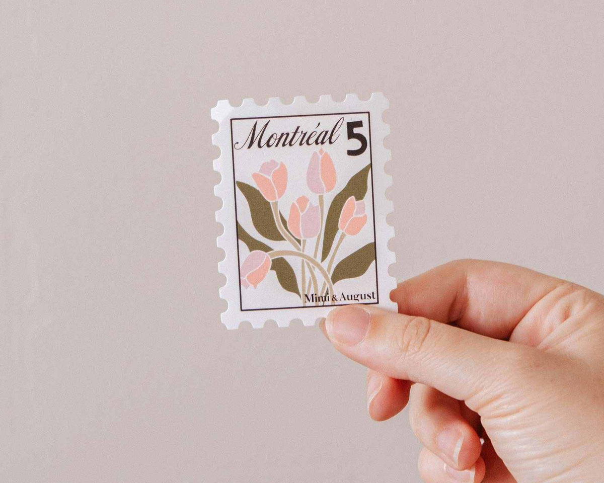 A hand holds the Montréal Stamp - Vinyl Sticker by Mimi & August, featuring pink flowers, green leaves, and "Montréal 5" at the top against a plain background.