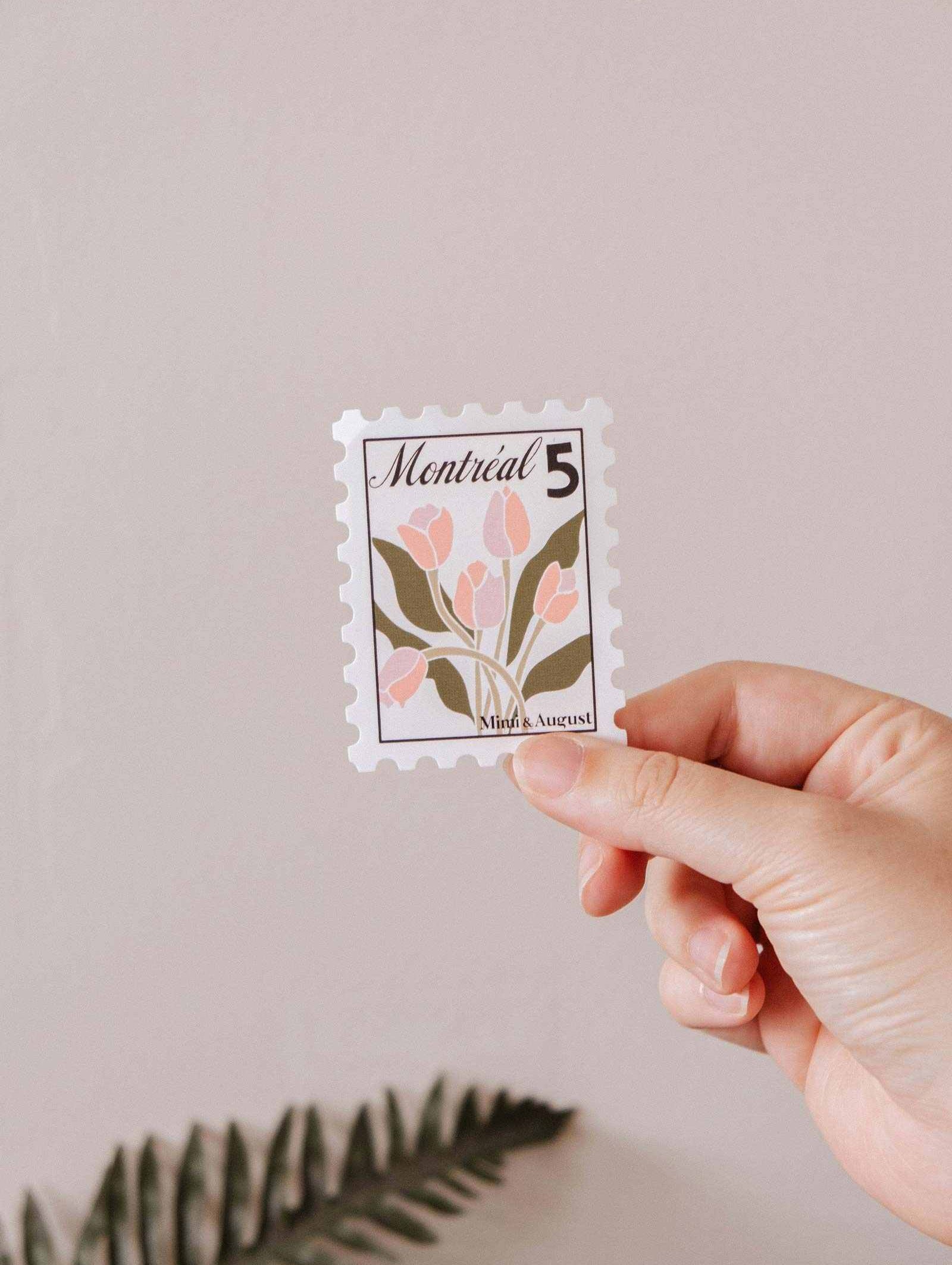 A hand holds the Montréal Stamp - Vinyl Sticker by Mimi & August, featuring pink flowers, green leaves, and "Montréal 5" at the top against a plain background.