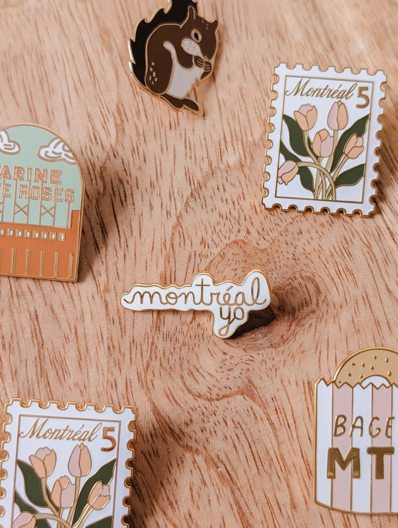 Five enamel pins from Mimi & August, including floral stamps, a squirrel, “Montréal Yo” enamel pin, “BAGEL MTL,” and “JARDIN DES ROSES,” are arranged on a wooden surface—each uniquely designed in Montreal.
