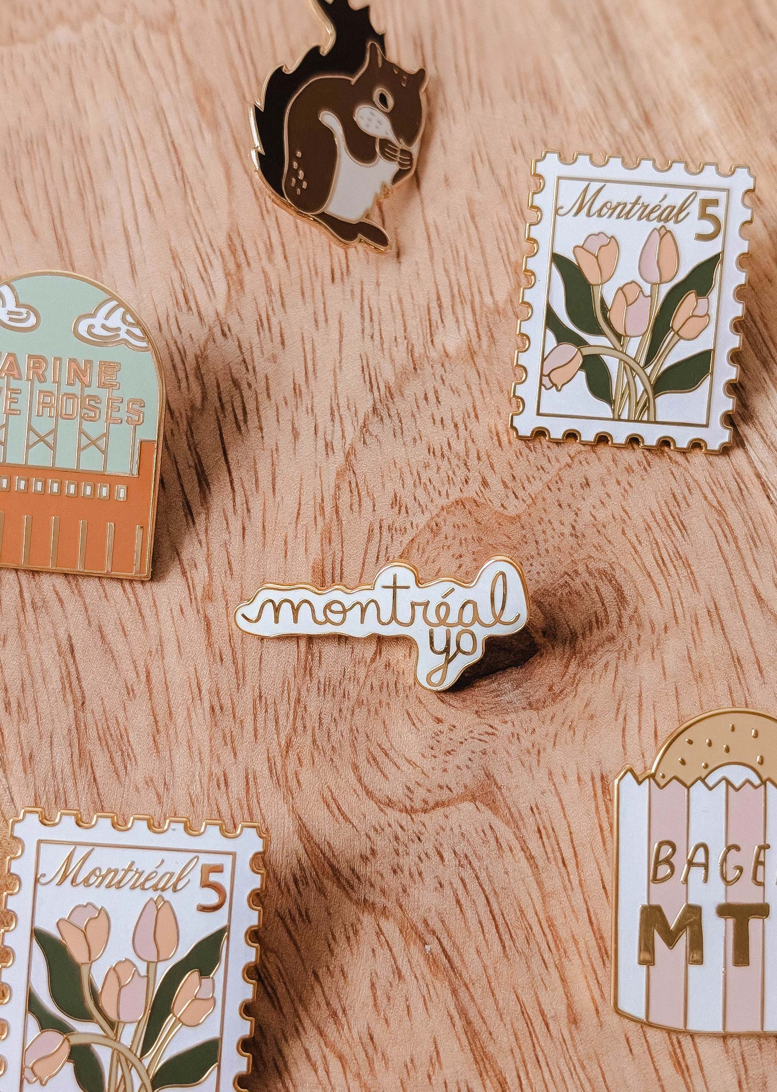 Five enamel pins from Mimi & August, including floral stamps, a squirrel, “Montréal Yo” enamel pin, “BAGEL MTL,” and “JARDIN DES ROSES,” are arranged on a wooden surface—each uniquely designed in Montreal.