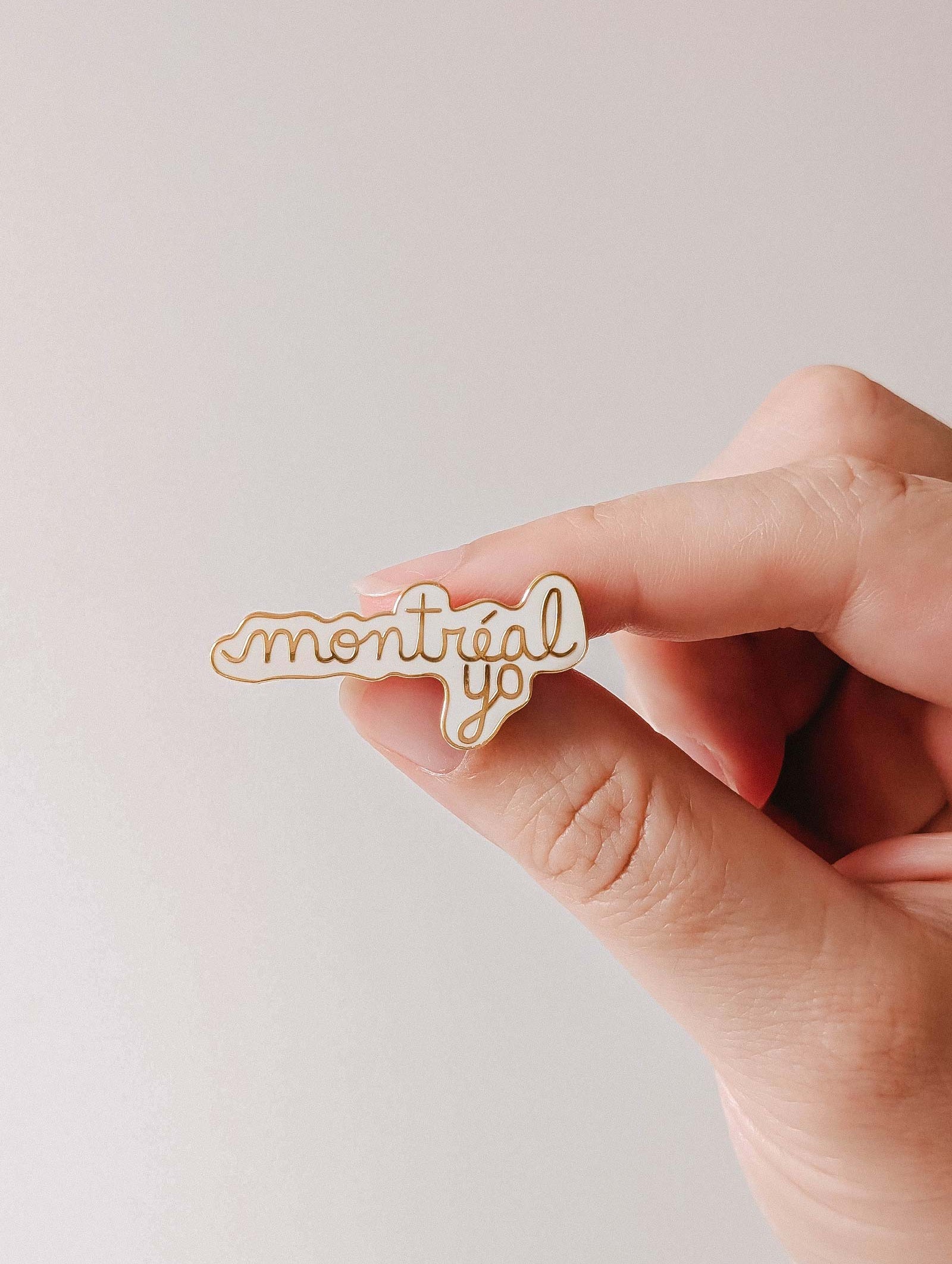 A hand holds the Montréal Yo Enamel Pin by Mimi & August, featuring "montréal yo" in cursive on a white background and crafted from gold tone brass enamel.