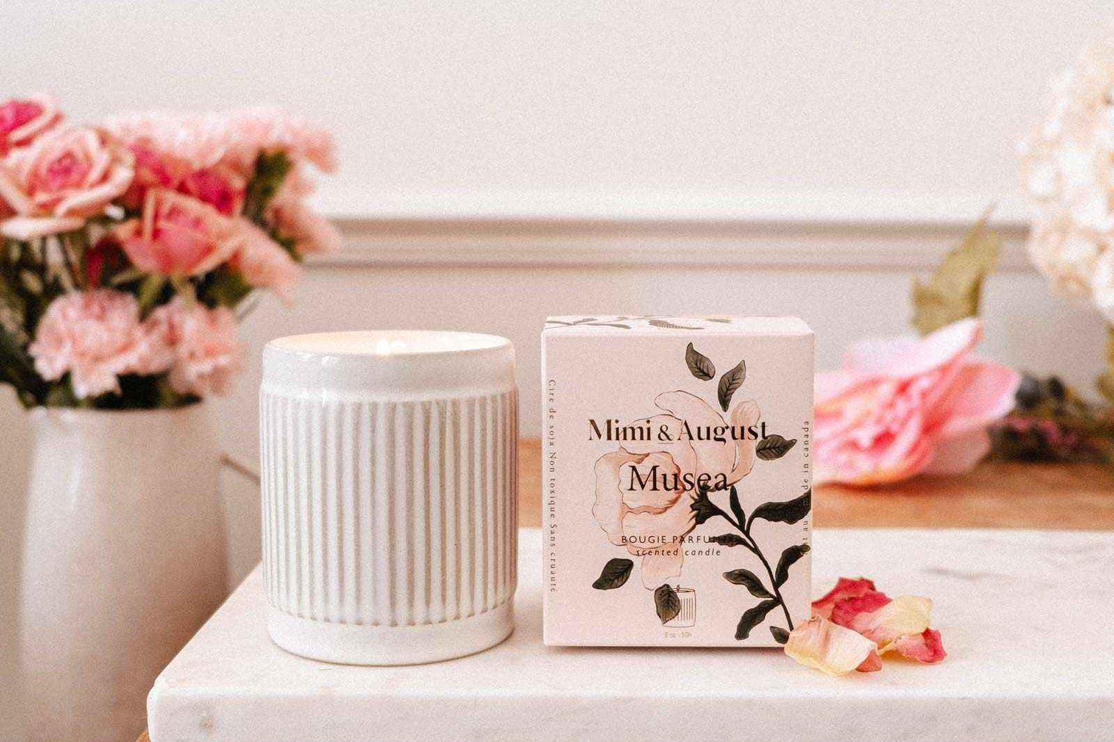 Une bougie de soya réutilisable Musea de Mimi & August est présentée sur une table en bois avec dessus en marbre, avec un vase de fleurs roses en arrière-plan.