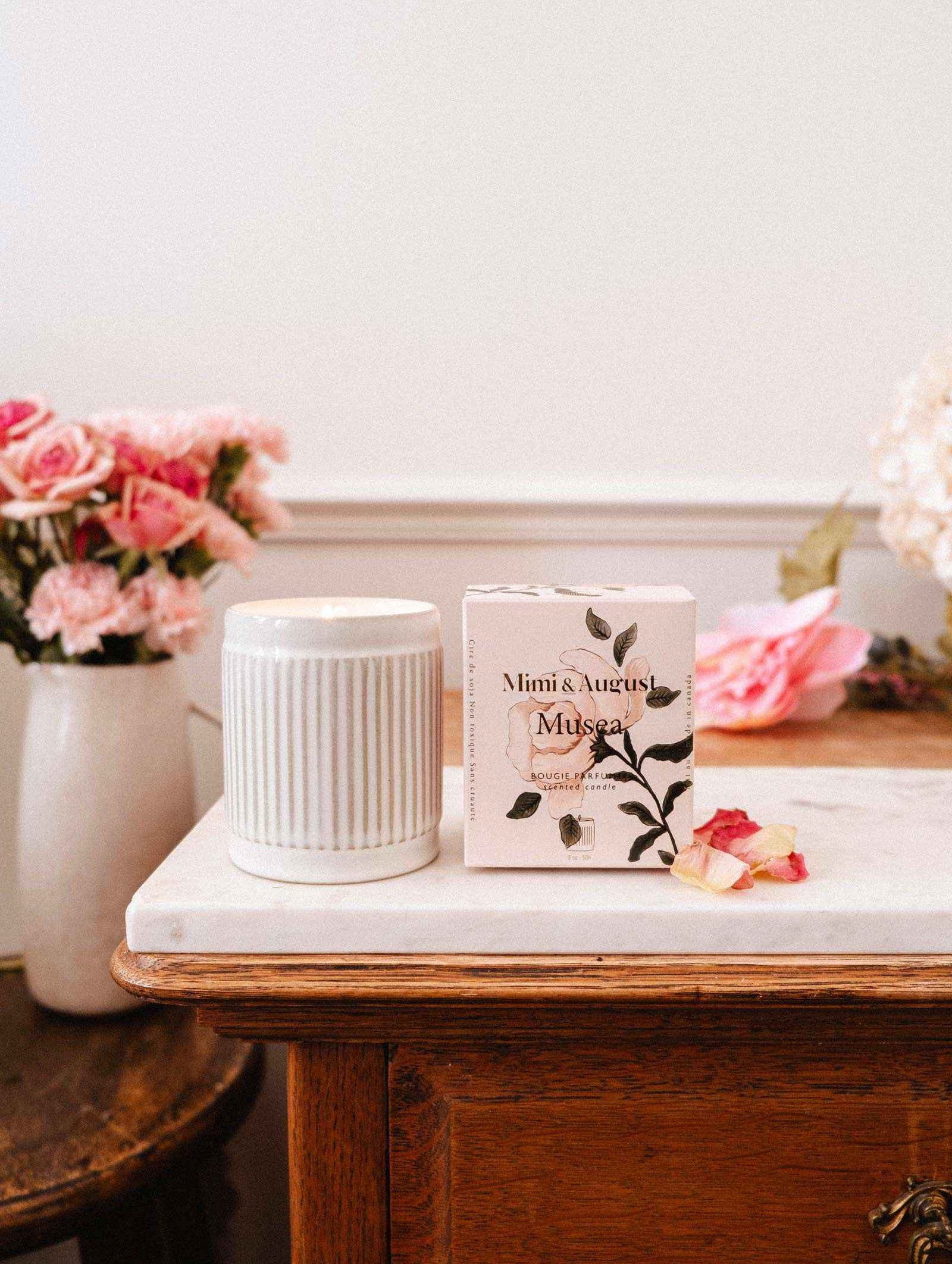Une bougie de soya réutilisable Musea de Mimi & August est présentée sur une table en bois avec dessus en marbre, avec un vase de fleurs roses en arrière-plan.
