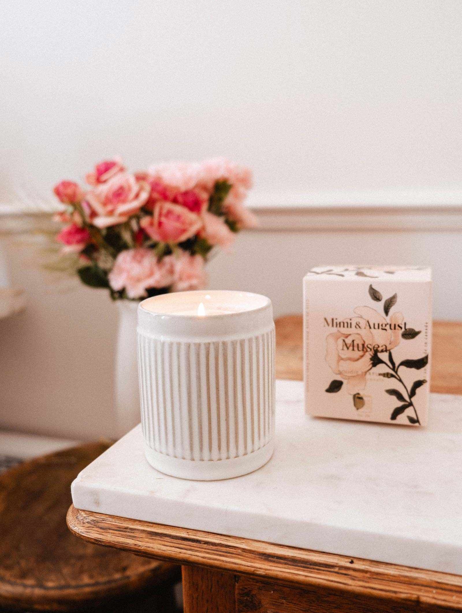La bougie de soya réutilisable Musea de Mimi & August, coulée à la main à Montréal, présente un design blanc cannelé et repose à côté de sa boîte florale sur du marbre avec des roses en arrière-plan — apportant élégance et chaleur à votre espace.