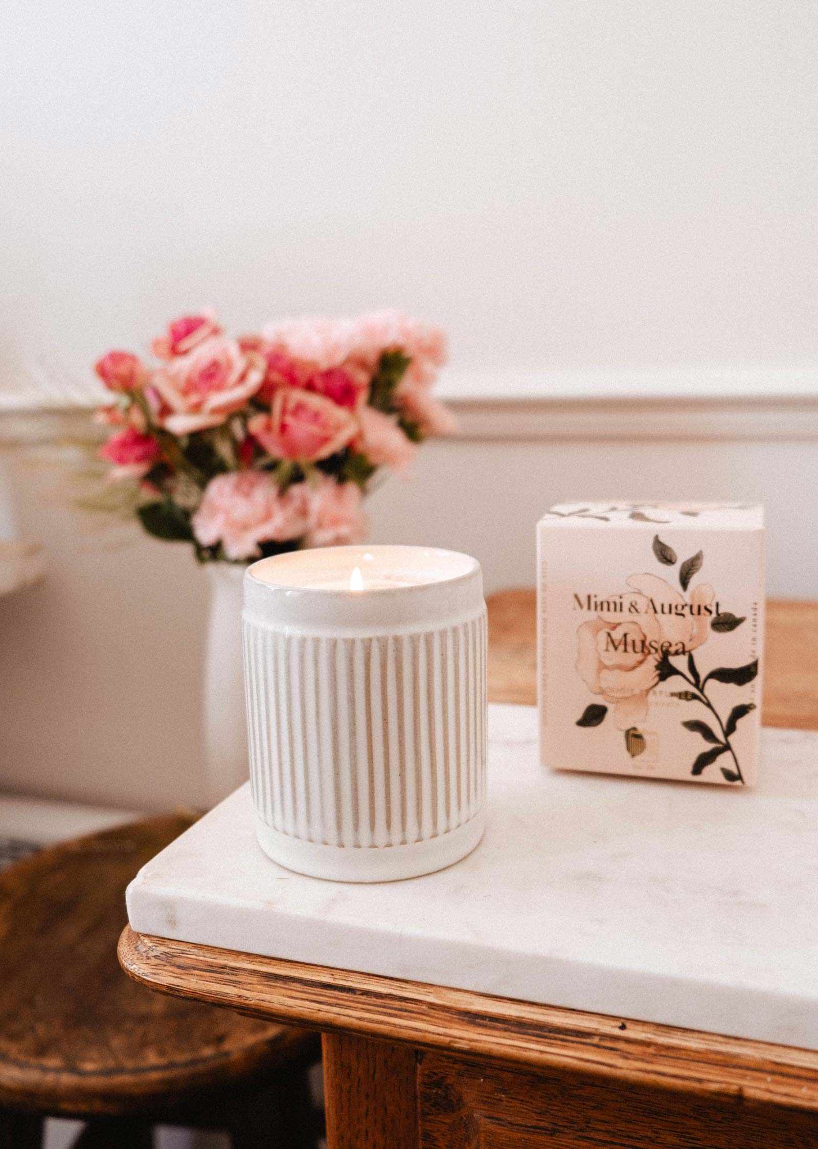 La bougie de soya réutilisable Musea de Mimi & August, coulée à la main à Montréal, présente un design blanc cannelé et repose à côté de sa boîte florale sur du marbre avec des roses en arrière-plan — apportant élégance et chaleur à votre espace.