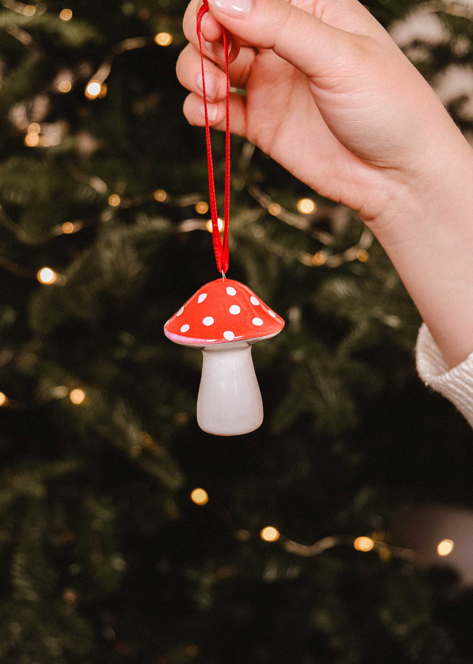 A hand holds the Mimi & August Mushroom Ceramic Christmas Ornament by a red ribbon before a glowing Christmas tree, bringing handcrafted holiday décor to the festive scene.