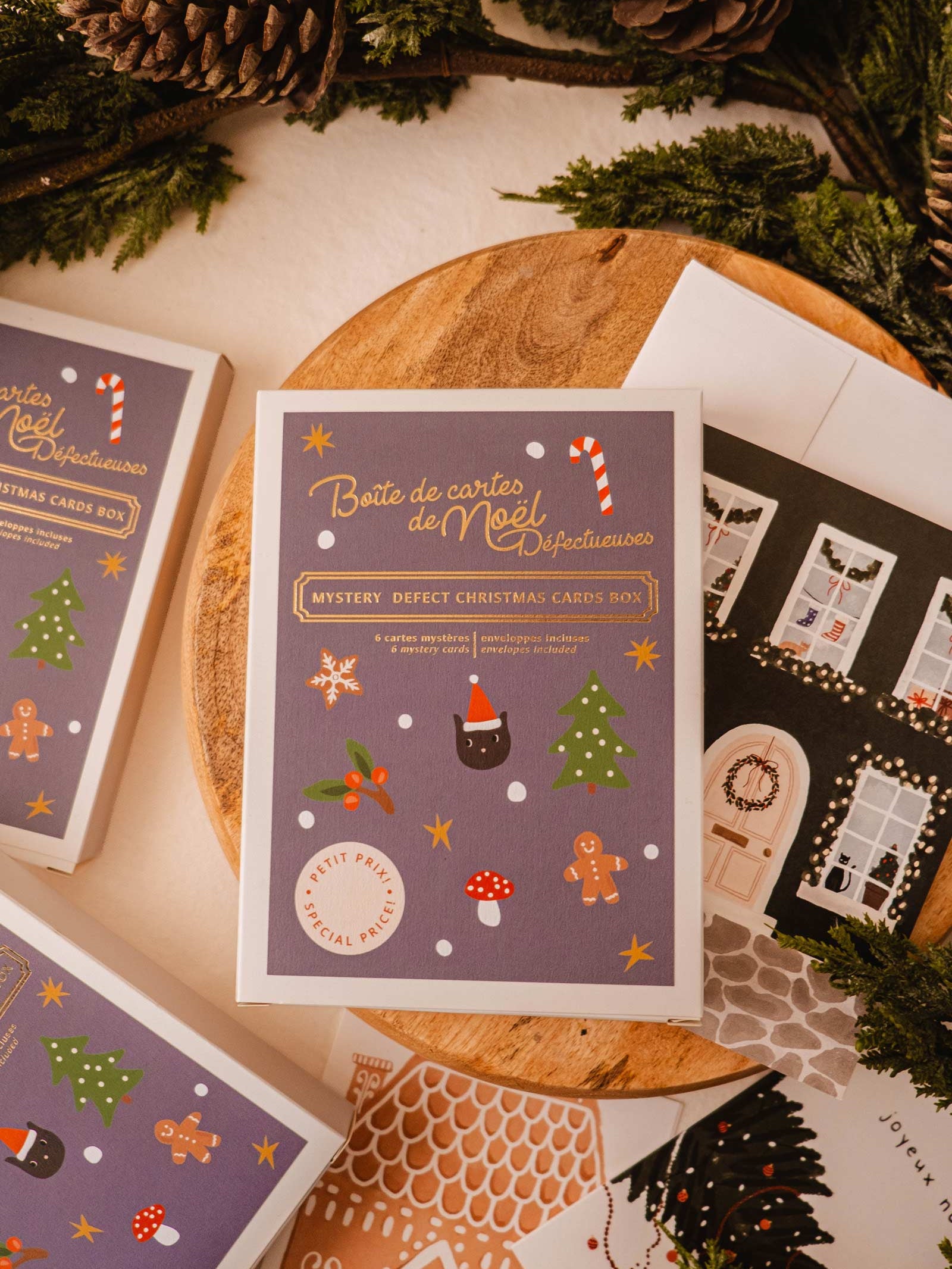 Embrace the holiday spirit with Mimi & August's Mystery Defect Christmas Card Box – Set of 6. This budget-friendly collection is elegantly adorned in purple, showcasing festive illustrations such as a cat in a Santa hat. It includes additional Christmas cards displayed on a wooden surface, surrounded by greenery.