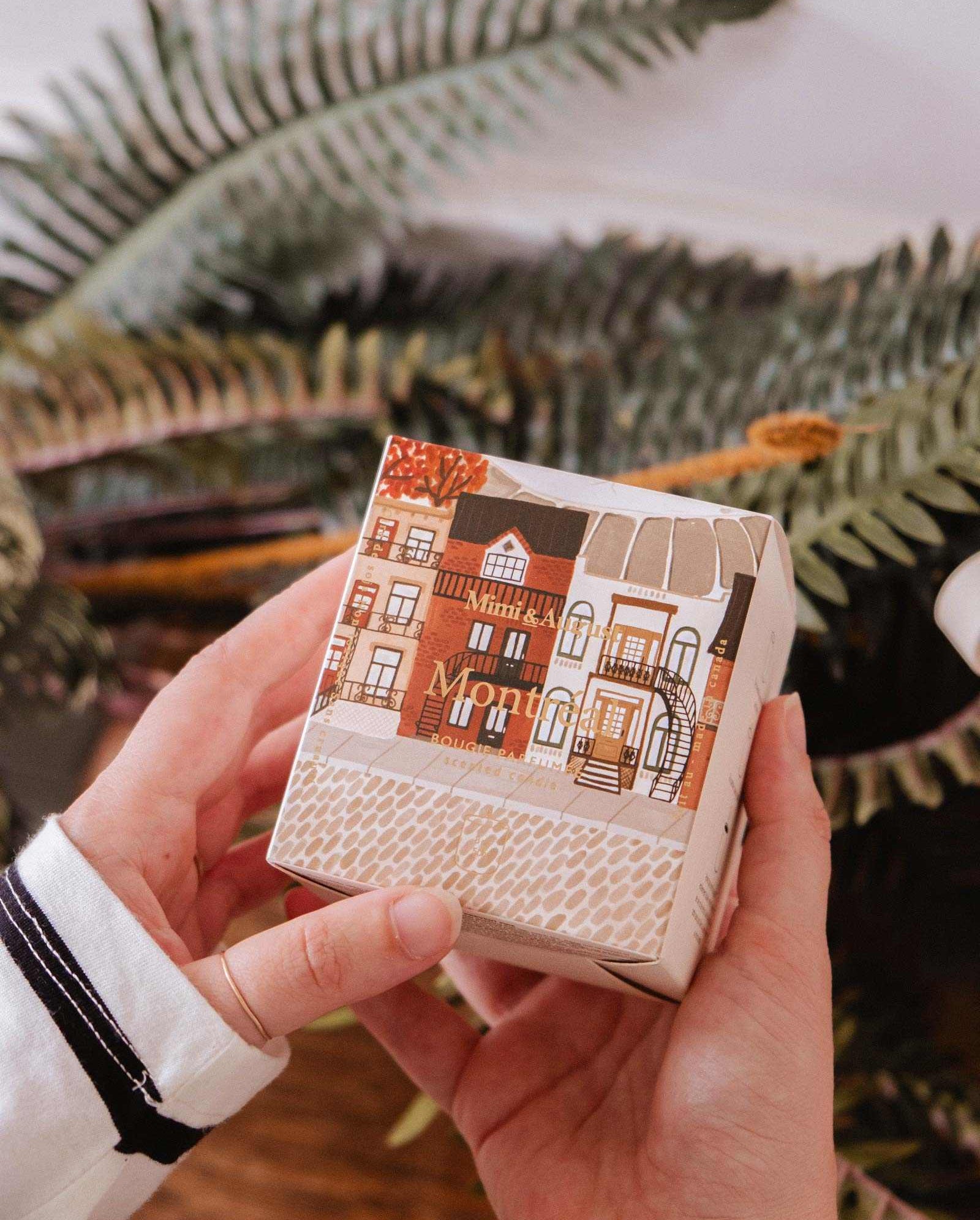 A person holds a Mimi & August Montreal - Reusable Candle box, featuring an illustrated cityscape, in front of green fern leaves.