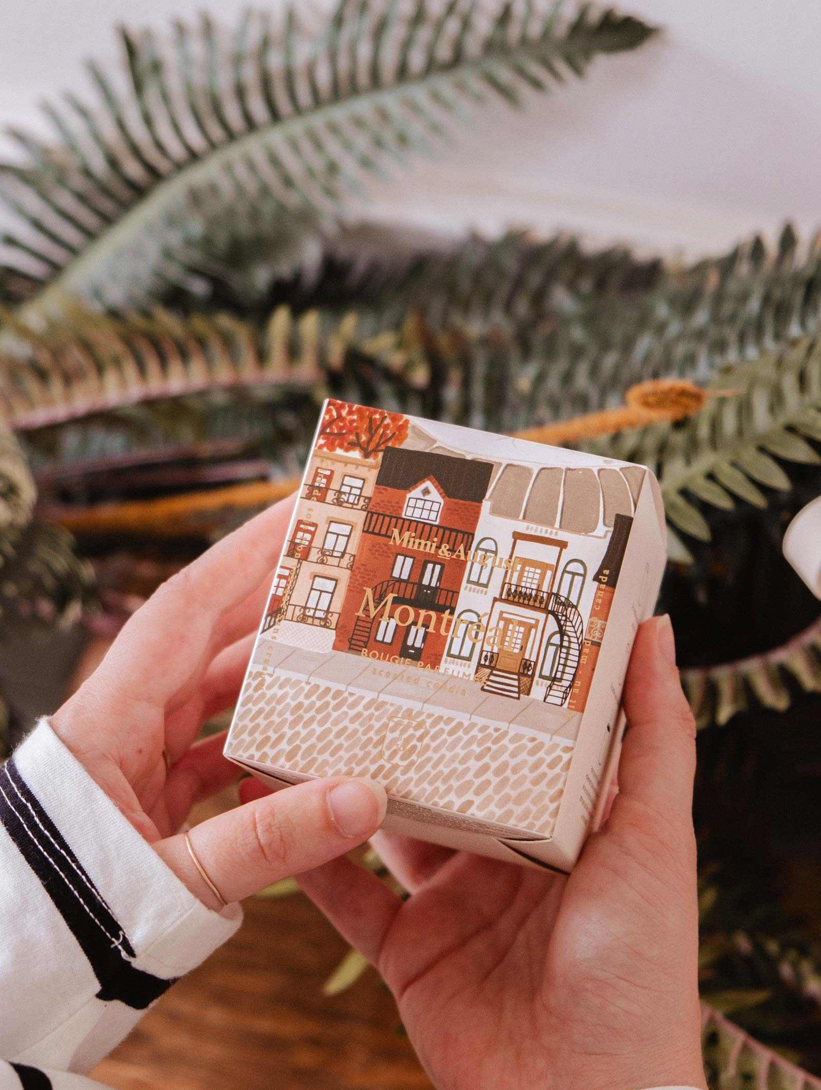 A person holds a Mimi & August Montreal - Reusable Candle box, featuring an illustrated cityscape, in front of green fern leaves.