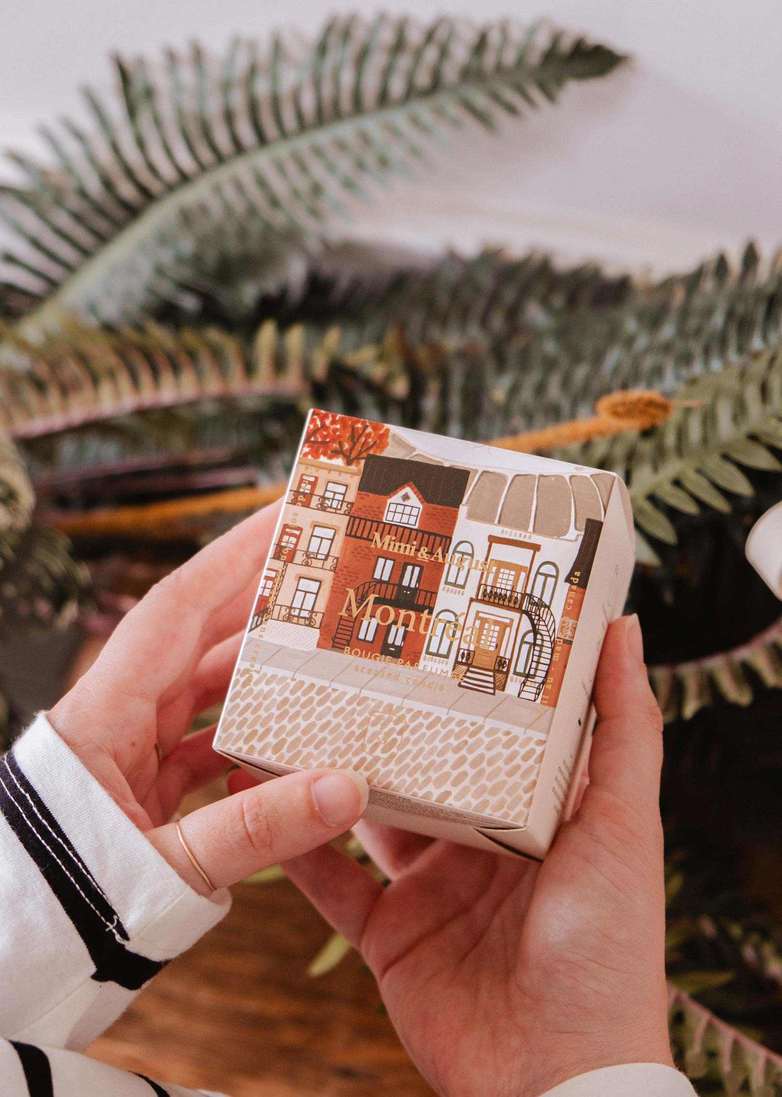 Une personne tient une boîte de la bougie réutilisable Montréal de Mimi & August, ornée d'un paysage urbain illustré, devant des feuilles de fougère vertes.