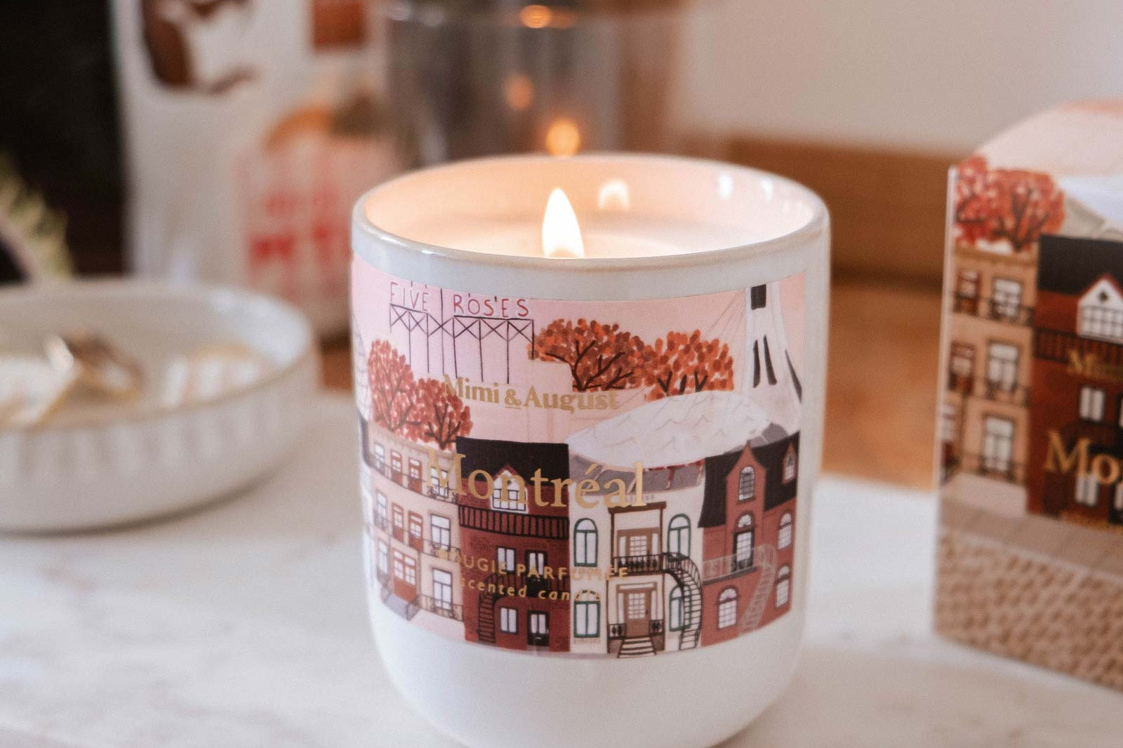 The Montreal - Reusable Candle by Mimi & August, a lit scented candle in a decorative white jar with illustrated buildings, sits on a marble tray atop a wooden surface, surrounded by small objects.