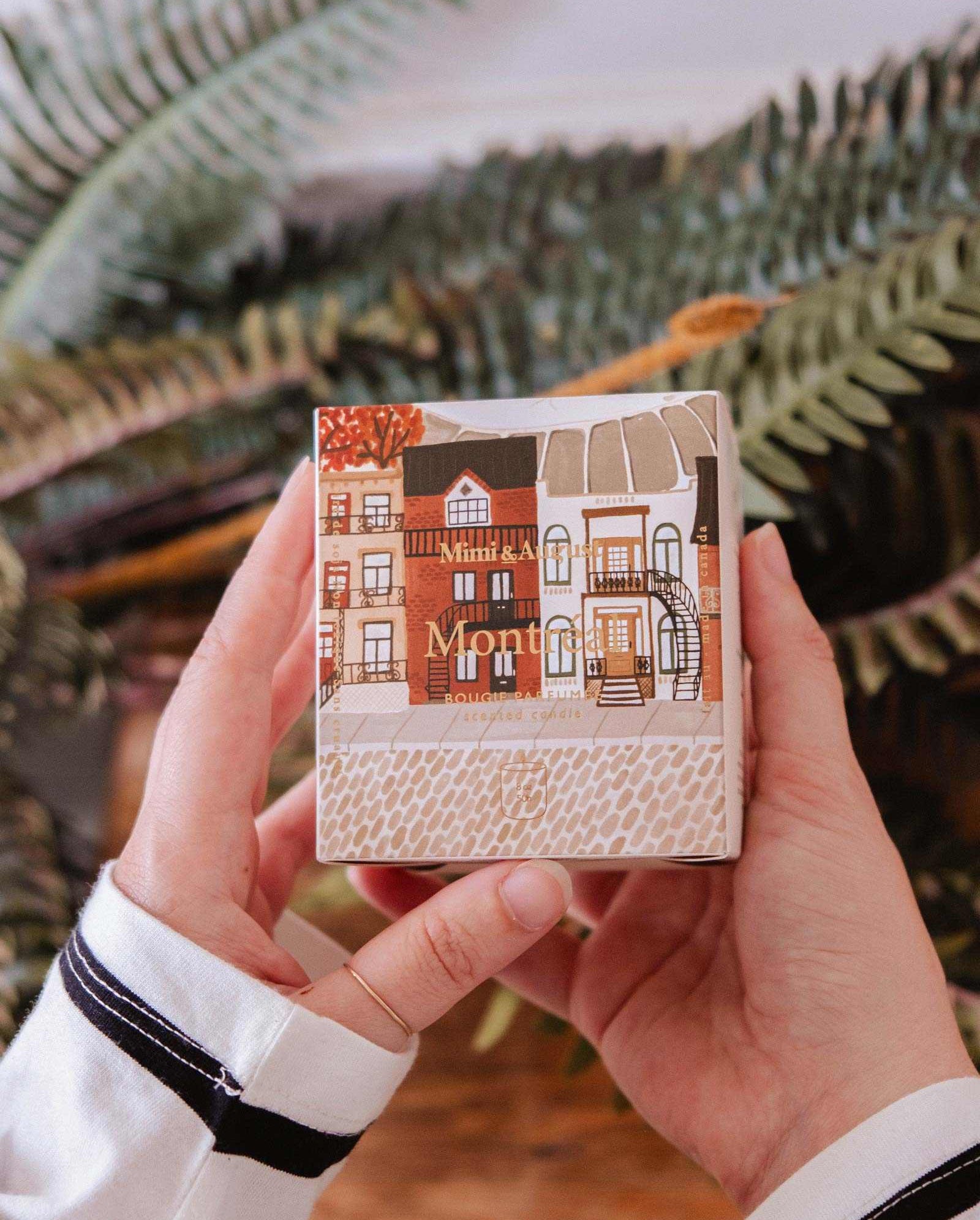 A person holds a small box featuring illustrated buildings, the packaging for Mimi & August’s Montreal - Reusable Candle, in front of green fern leaves.