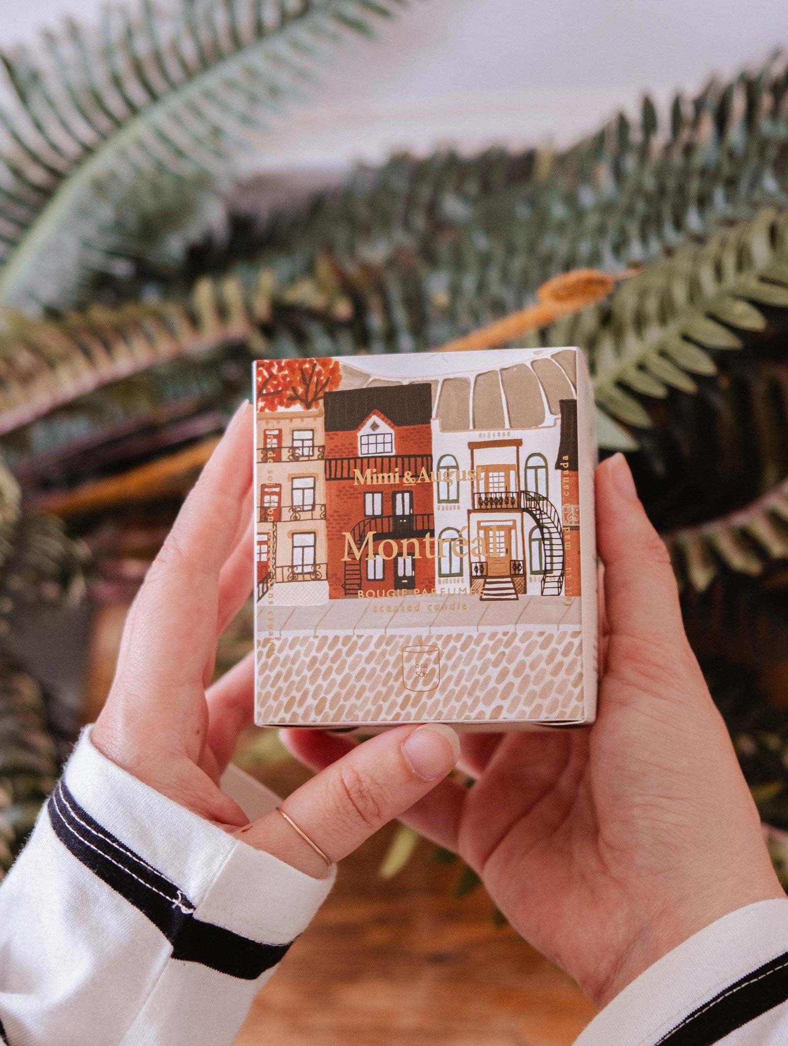 A person holds a small box featuring illustrated buildings, the packaging for Mimi & August’s Montreal - Reusable Candle, in front of green fern leaves.