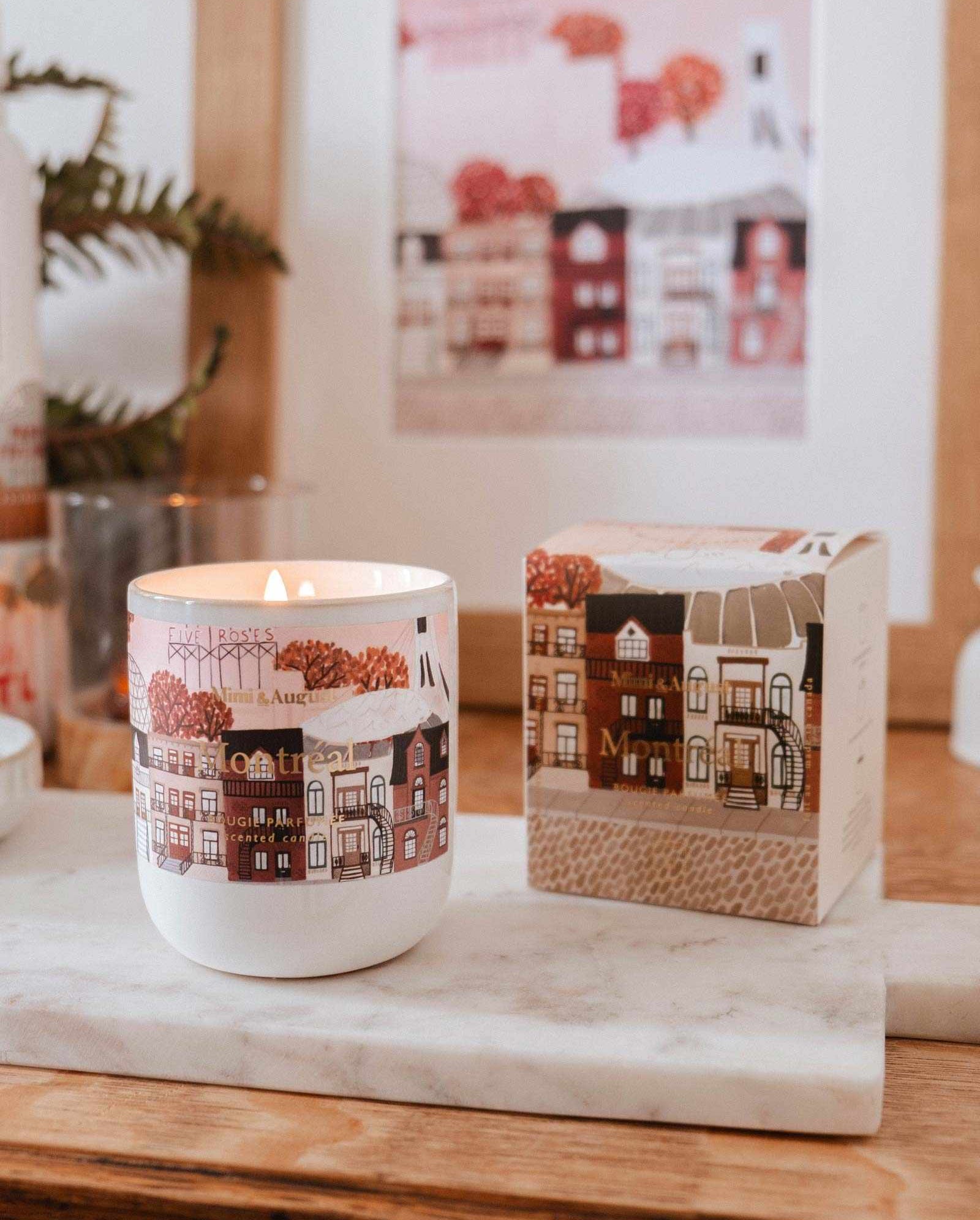 The Montreal - Reusable Candle by Mimi & August, featuring illustrated houses on its container, sits on a marble tray beside its matching box with a framed print in the background.