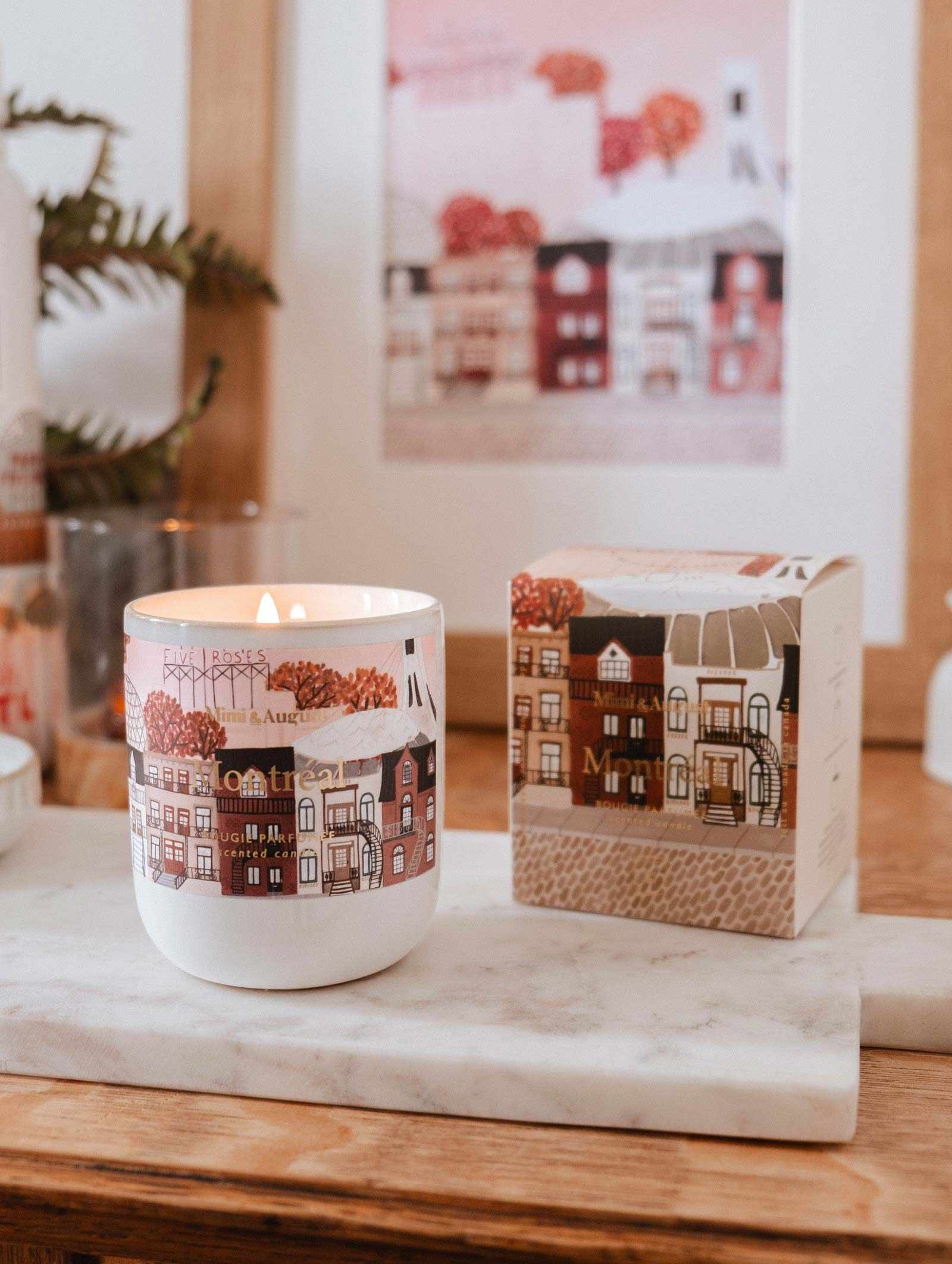 The Montreal - Reusable Candle by Mimi & August, featuring illustrated houses on its container, sits on a marble tray beside its matching box with a framed print in the background.
