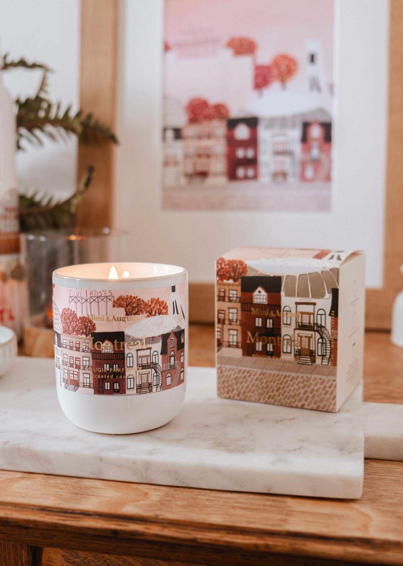 La bougie réutilisable Montréal de Mimi & August, ornée de maisons illustrées sur son contenant, repose sur un plateau en marbre à côté de sa boîte assortie avec une affiche encadrée en arrière-plan.