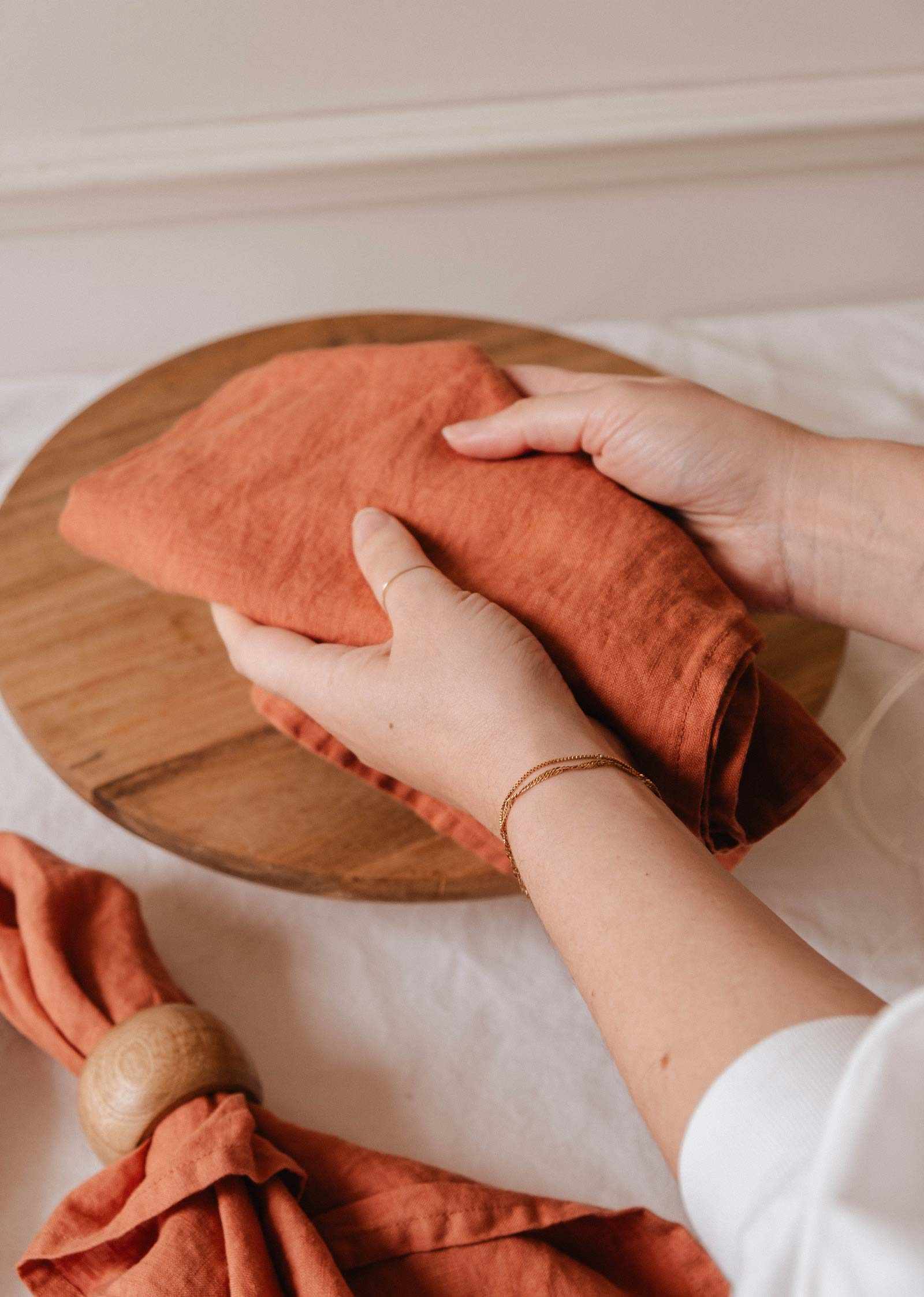 A person folds a Mimi & August Tea Towel Soft Linen, resembling a napkin, over a round wooden board, with another tea towel and napkin ring resting on a white tablecloth nearby.
