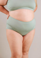 A close-up of a woman wearing the Mimi & August Paloma Agave High Waist Bikini Bottom, showing her midsection and thighs against a neutral background.