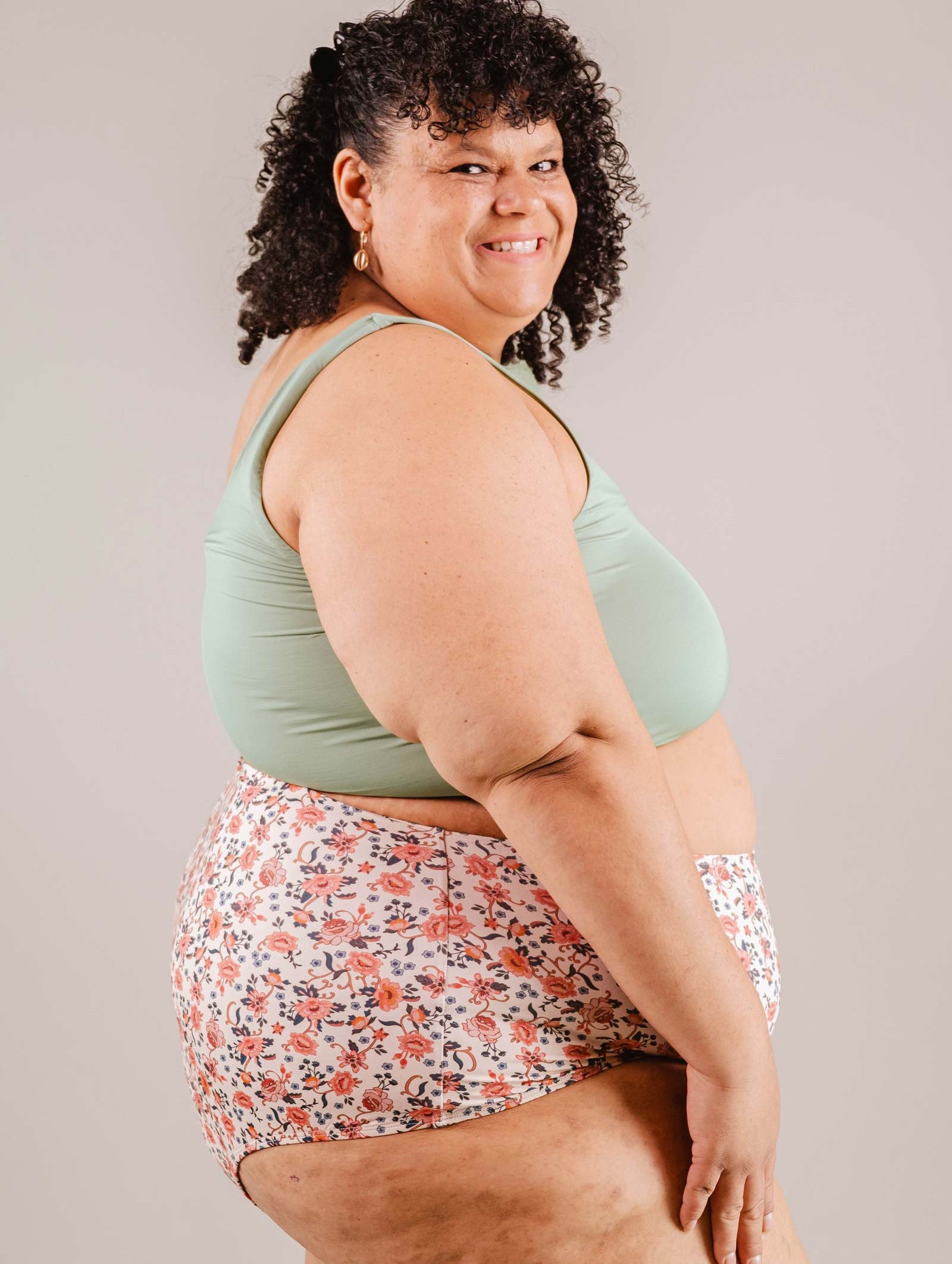 A woman with curly hair smiling at the camera, wearing a mint green tank top and Mimi & August's Paloma Amour High Waist Bikini Bottom, standing in a neutral-colored studio.