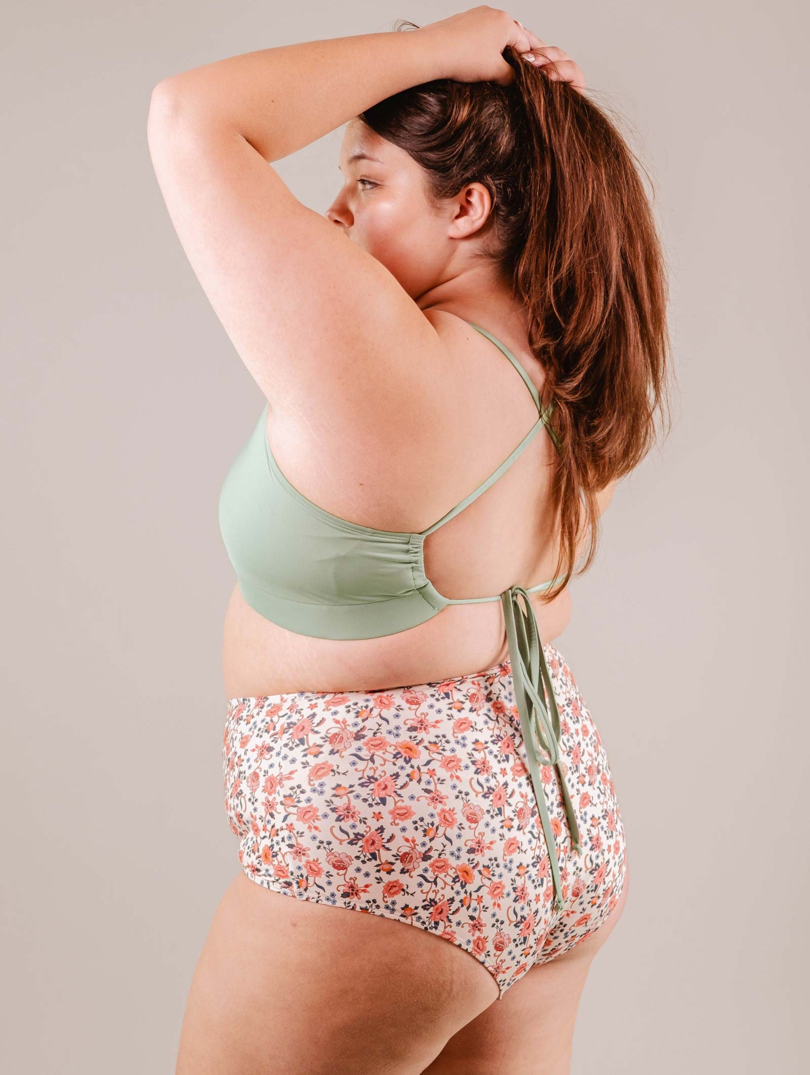 A woman in a Paloma Amour high waist bikini bottom from Mimi & August and floral shorts poses with her arm raised, adjusting her hair.