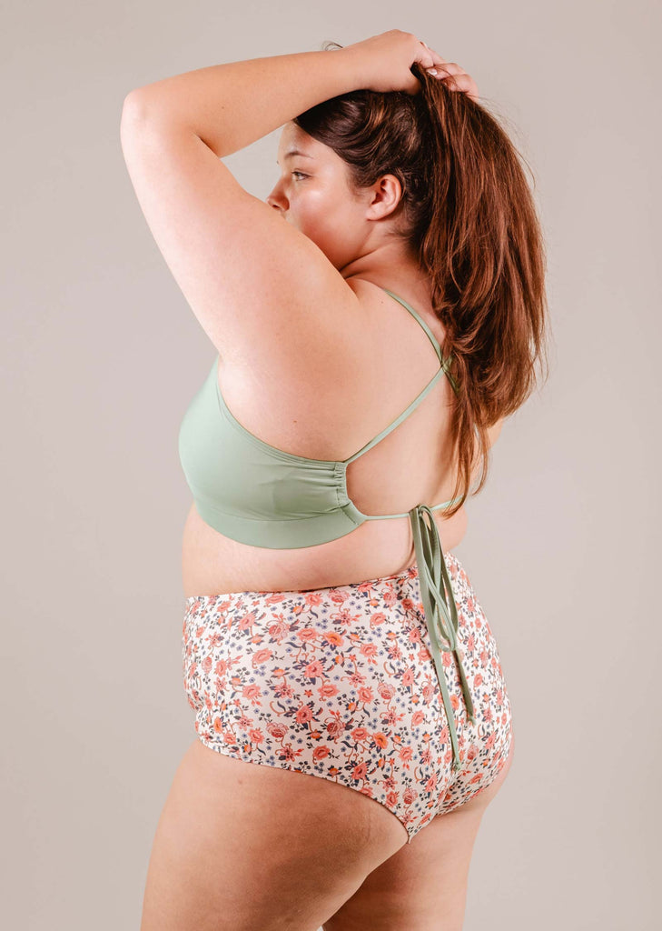A woman in a Paloma Amour high waist bikini bottom from Mimi & August and floral shorts poses with her arm raised, adjusting her hair.