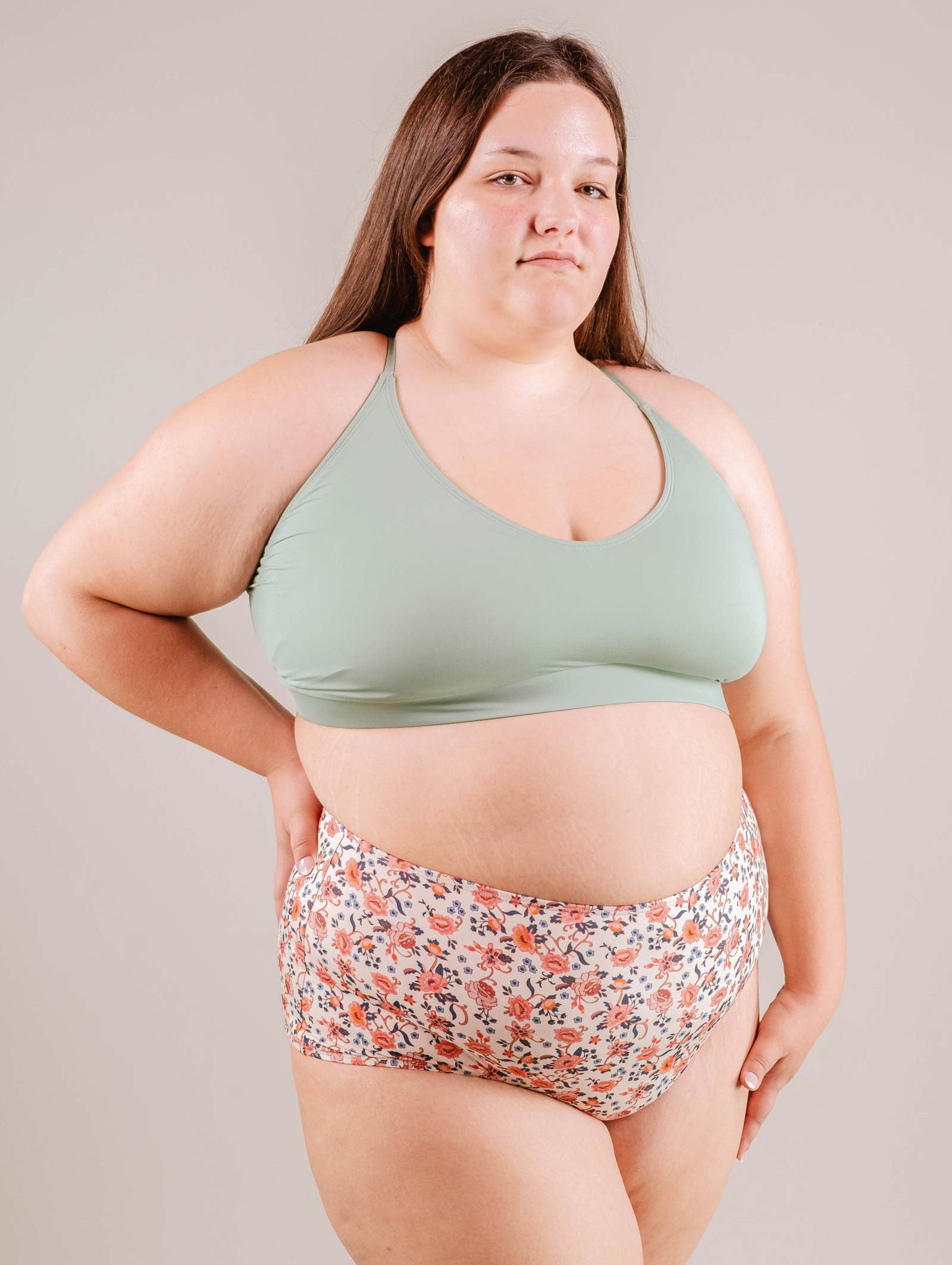 A confident plus-size woman in a green sports bra and Paloma Amour High Waist Bikini Bottom shorts standing against a neutral background by Mimi & August.