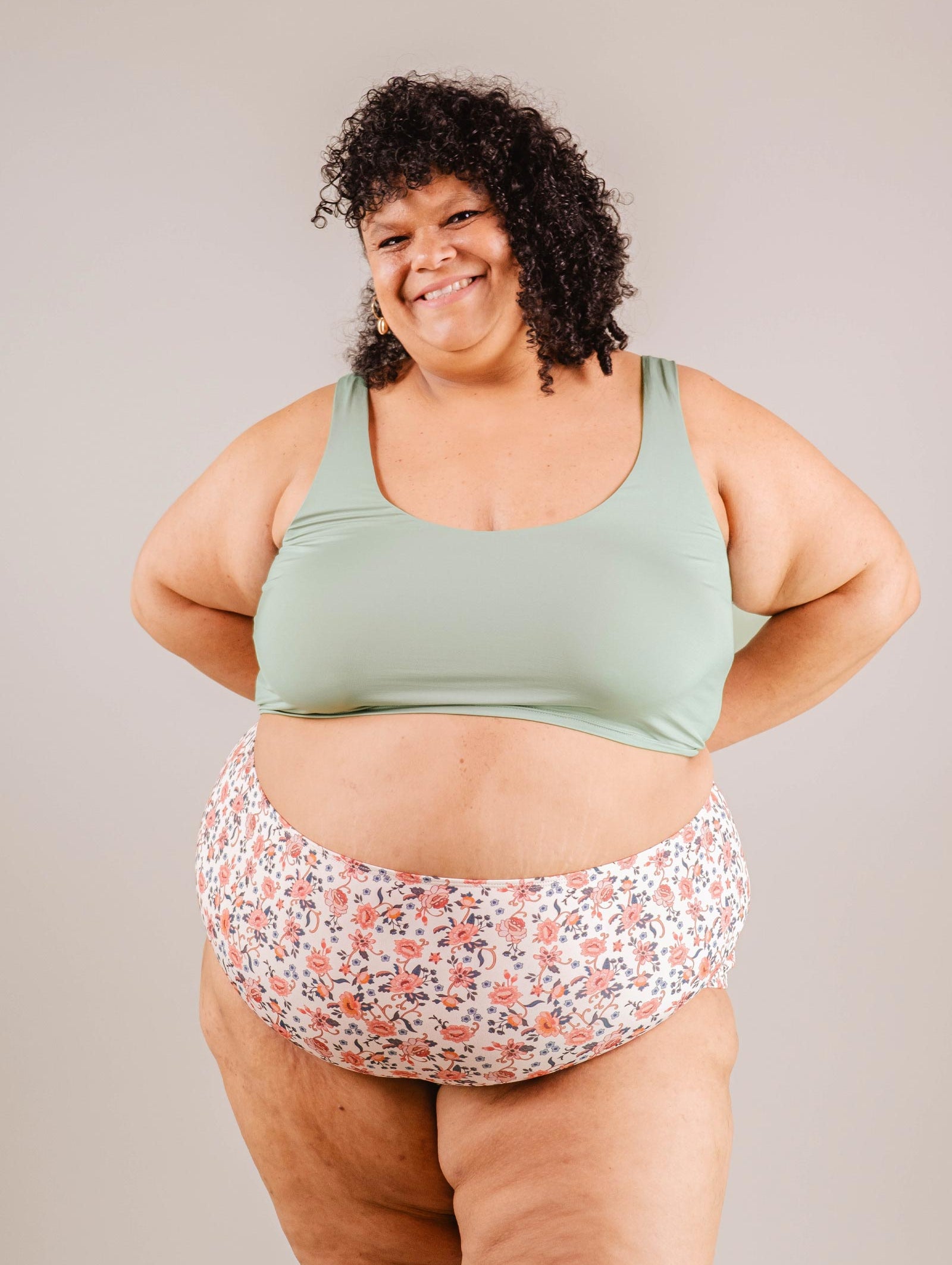 A cheerful woman in a green sports bra and Paloma Amour High Waist Bikini Bottom by Mimi & August, standing confidently with hands on hips against a neutral background.