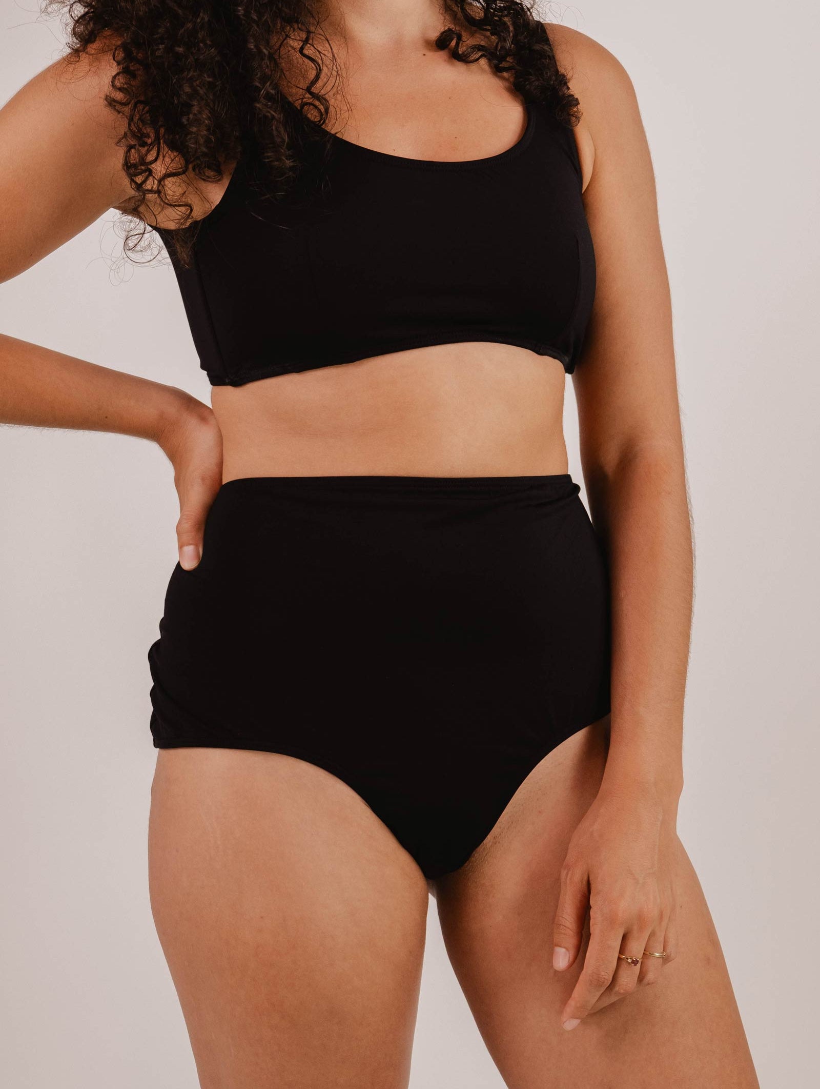 Person wearing a black two-piece swimsuit, featuring the Paloma Black High Waist Bikini Bottom by Mimi & August, with one hand resting on their hip against a plain background.