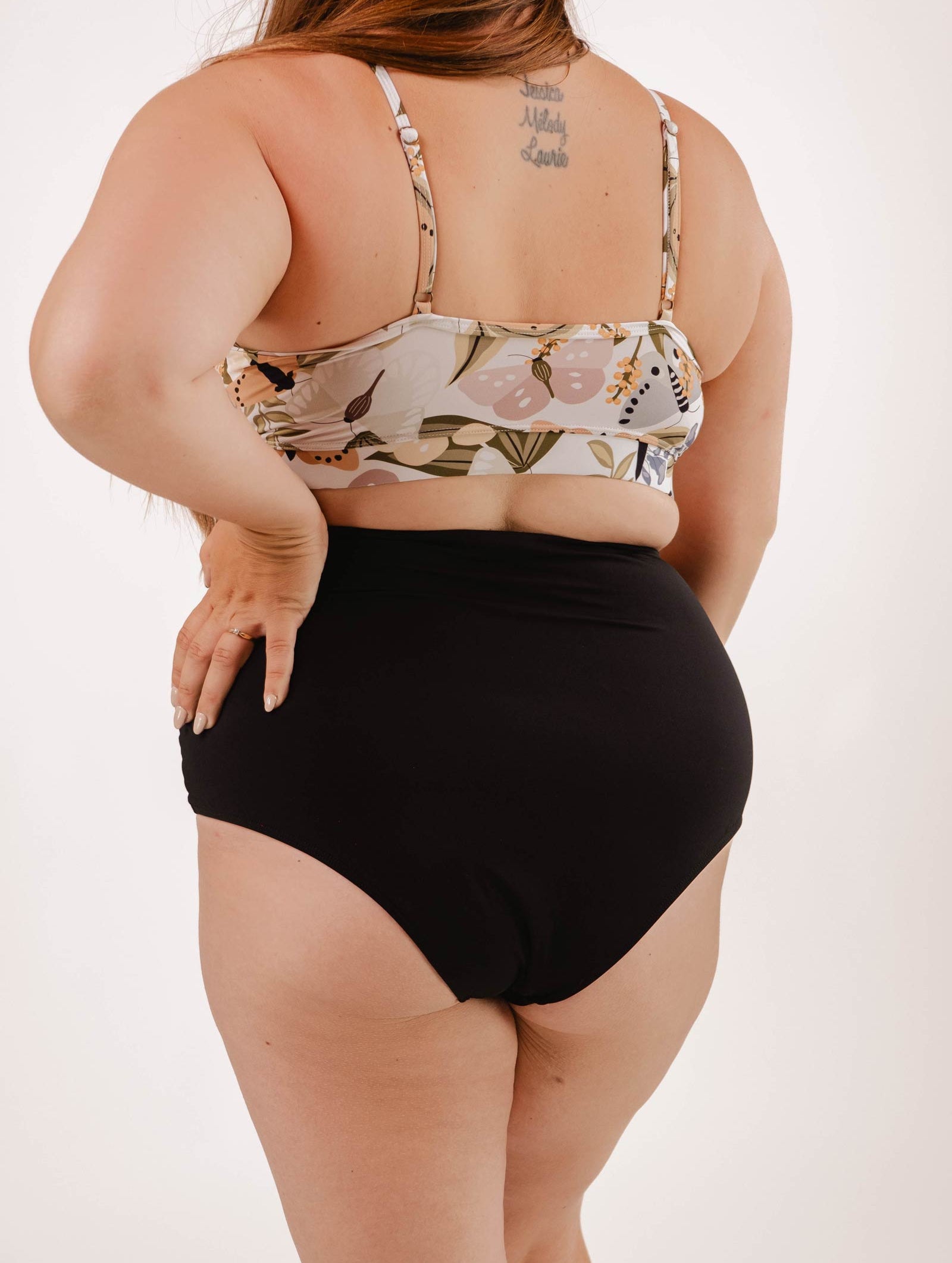A person is standing with their back to the camera, wearing a floral bikini top and Mimi & August Paloma Black High Waist Bikini Bottom, a true beachwear staple, with a tattoo on the upper back.