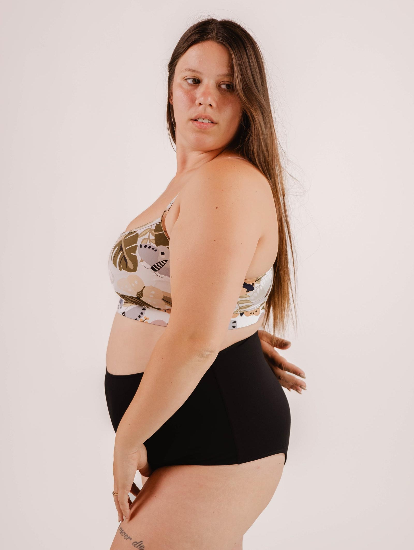 A woman with long hair stands sideways, wearing a patterned sports bra and Mimi & August Paloma Black High Waist Bikini Bottom against a plain background.