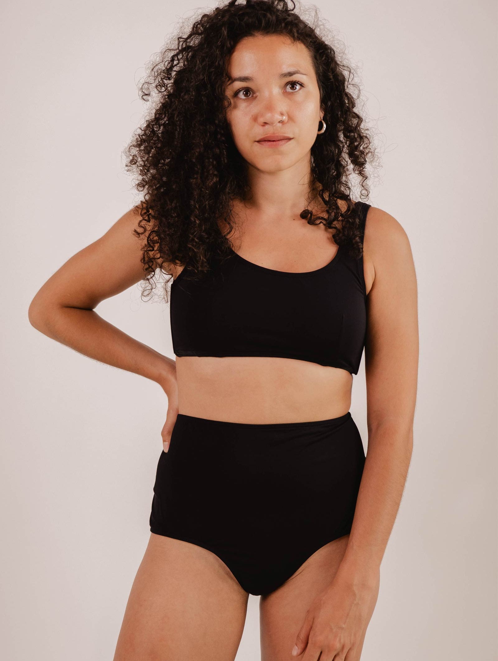 A woman with curly hair is wearing a black two-piece swimsuit. She is standing with one hand on her hip, looking slightly off camera against a plain background. The high-waisted swimwear style of her Paloma Black High Waist Bikini Bottom from Mimi & August exudes timeless elegance and confidence.